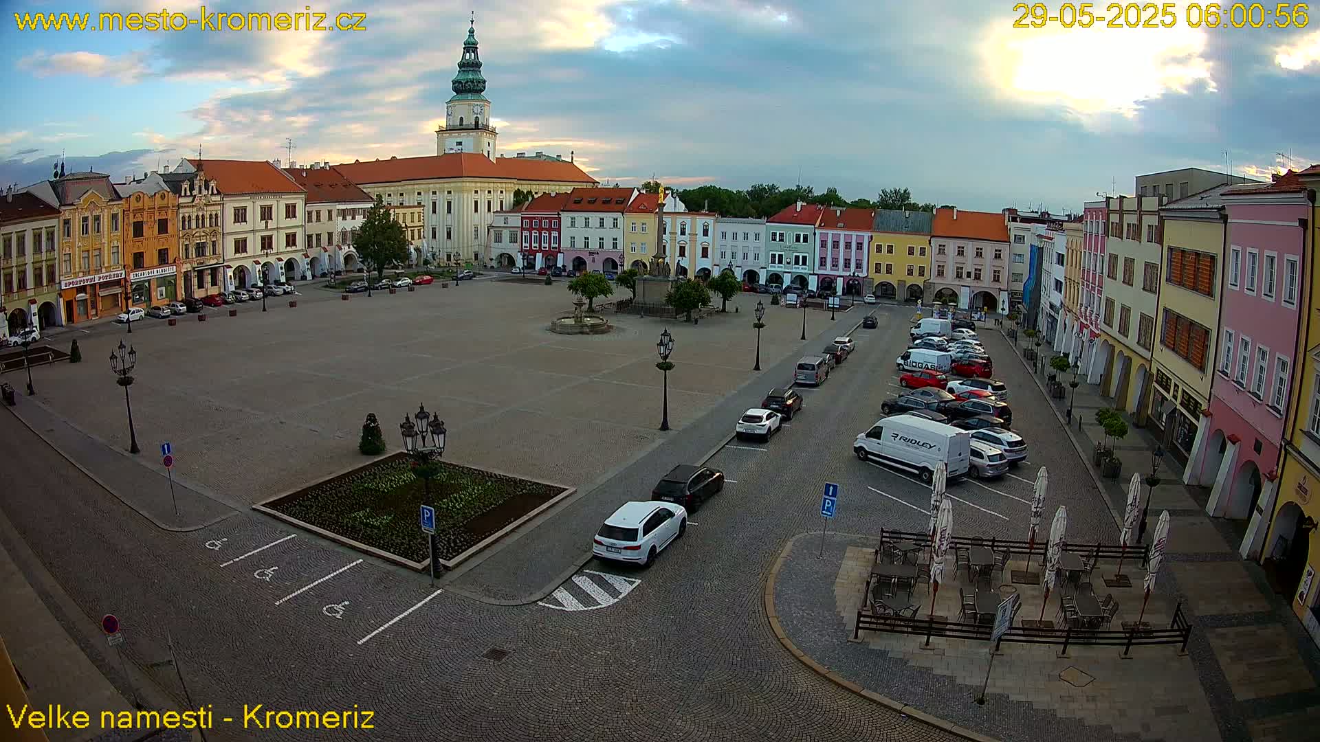 A large town square with colorful buildings surrounding it, under partly cloudy skies, features several cars parked along the edges, and outdoor seating in the lower right corner.