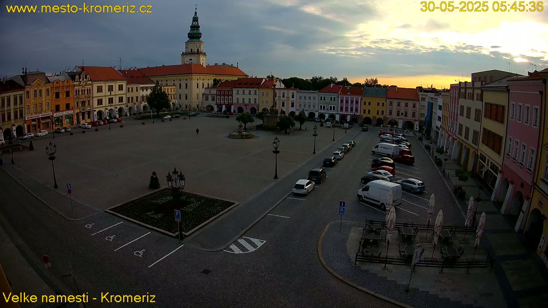 An aerial view of a town square at sunrise, with colorful buildings surrounding a large paved area containing parked cars and a few people.