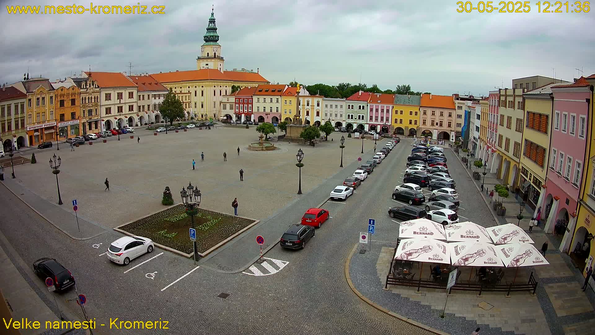 A large town square with colorful buildings surrounding it, under an overcast sky, is partially filled with parked cars and a few pedestrians.