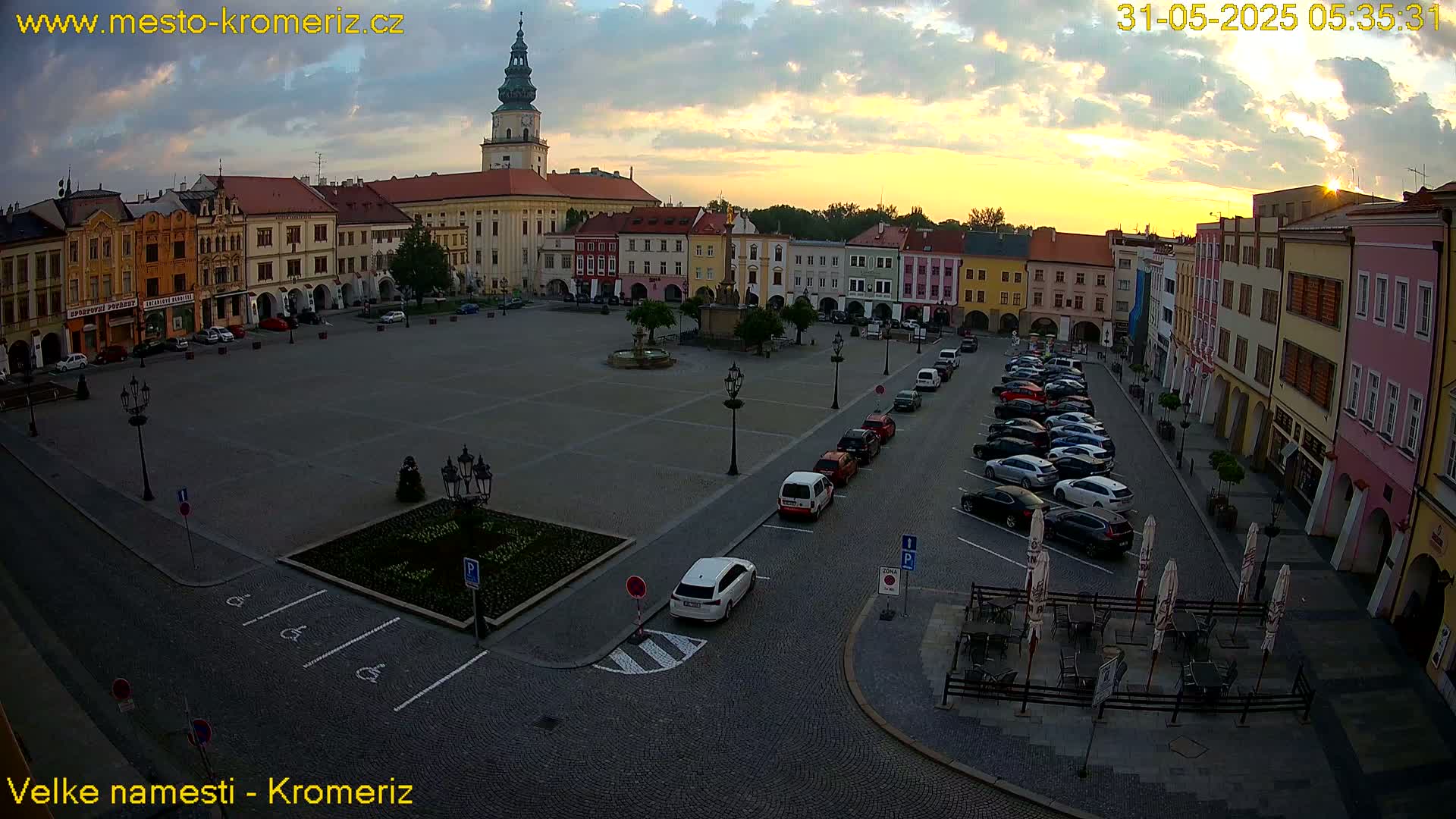 A large town square with colorful buildings surrounding it, cars parked along the sides, and a few outdoor tables and umbrellas set up, all under a partly cloudy sunrise sky.