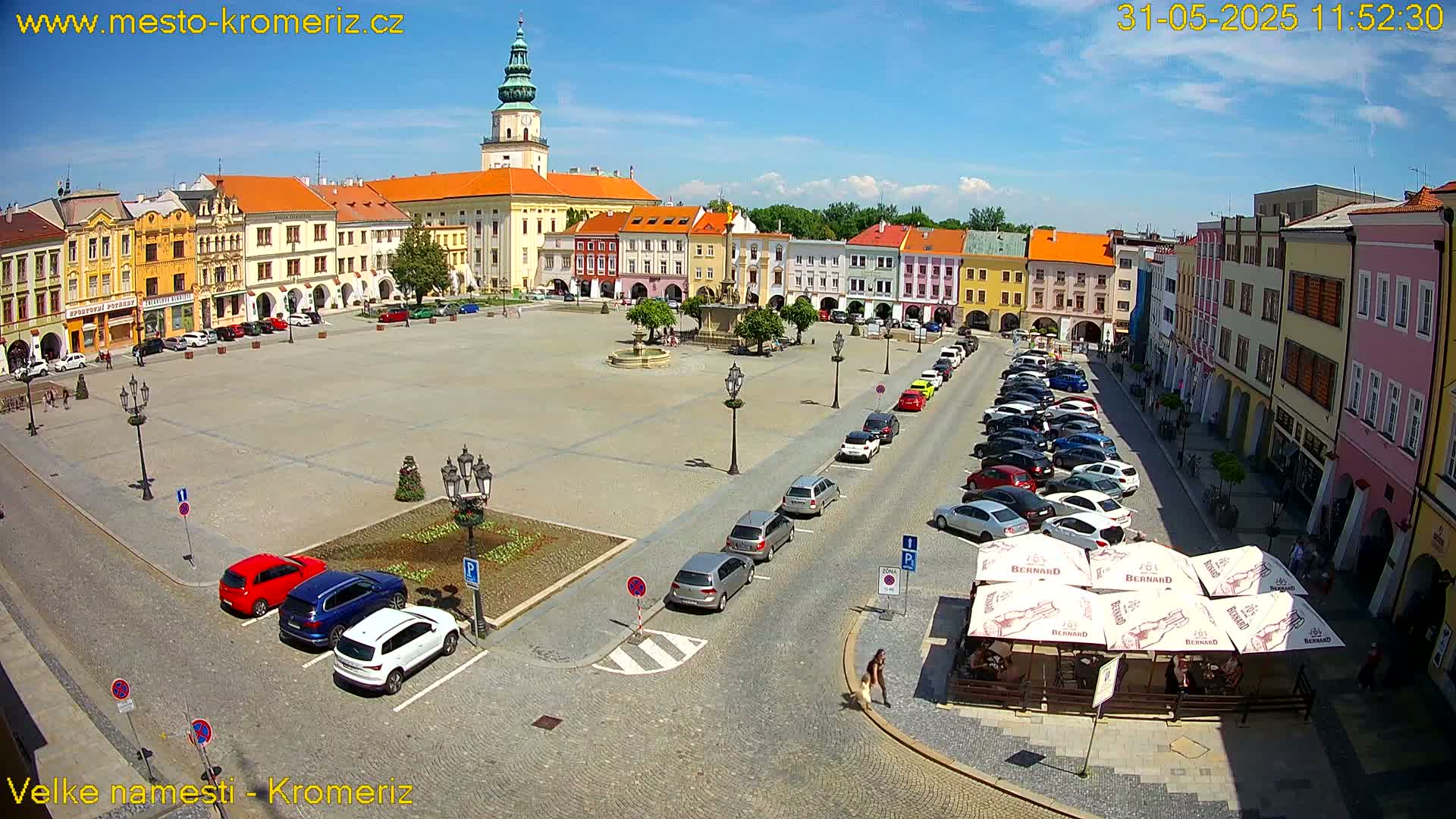 A sunny town square with colorful buildings, a fountain, parked cars, and outdoor seating at a cafe.