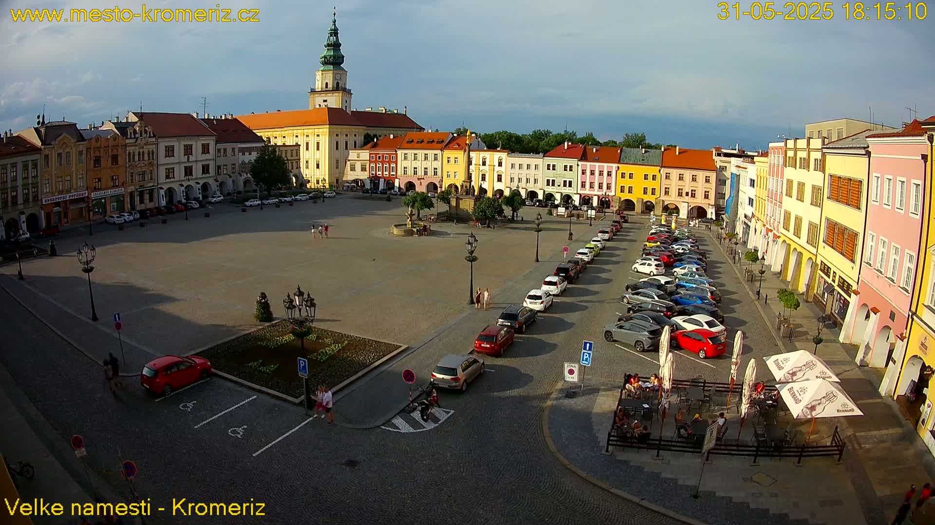 A large town square with colorful buildings surrounding it, under a partly cloudy sky, shows several cars parked along the sides and a few people walking around.