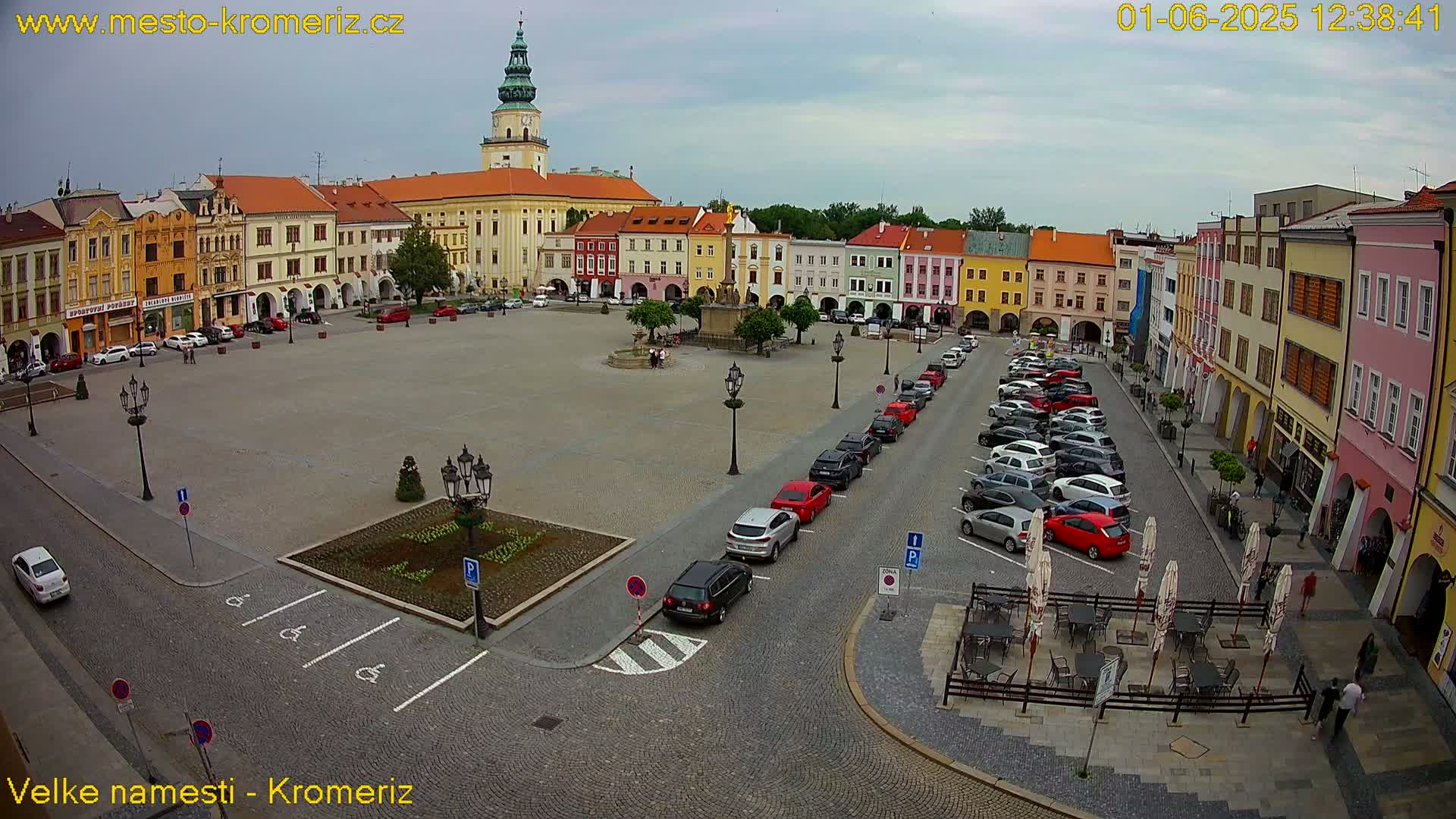 A large town square with colorful buildings surrounding it, under an overcast sky, is filled with parked cars and a few pedestrians.