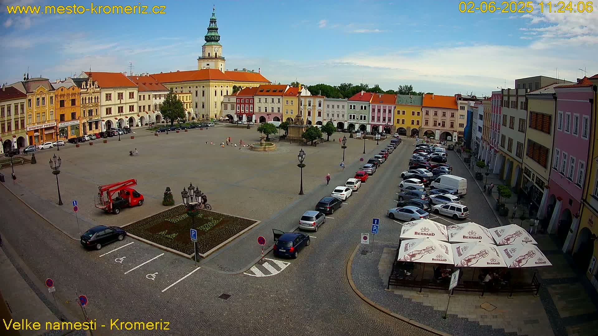 A large town square with colorful buildings, a fountain, and parked cars under a mostly clear sky.