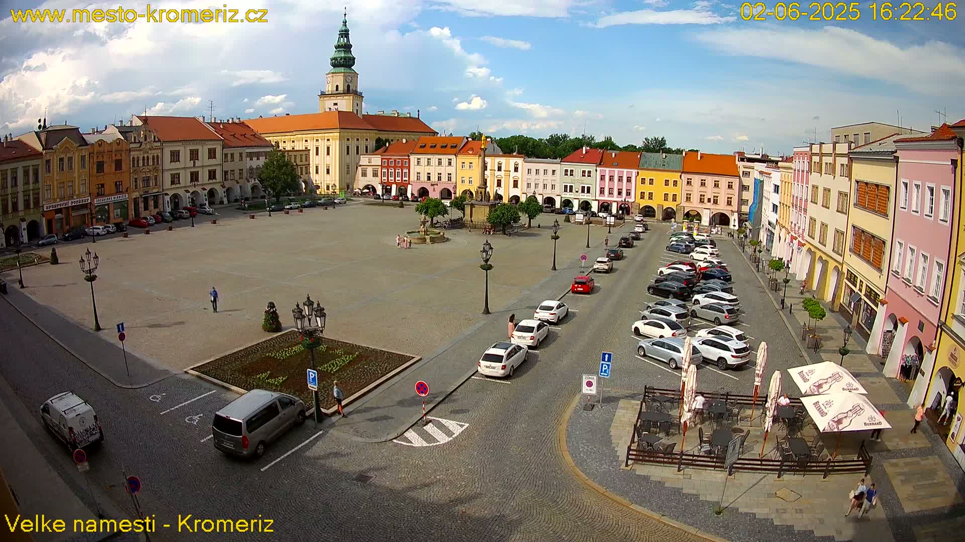 A large town square with colorful buildings surrounding it, under a partly cloudy sky, contains parked cars and several people walking around.