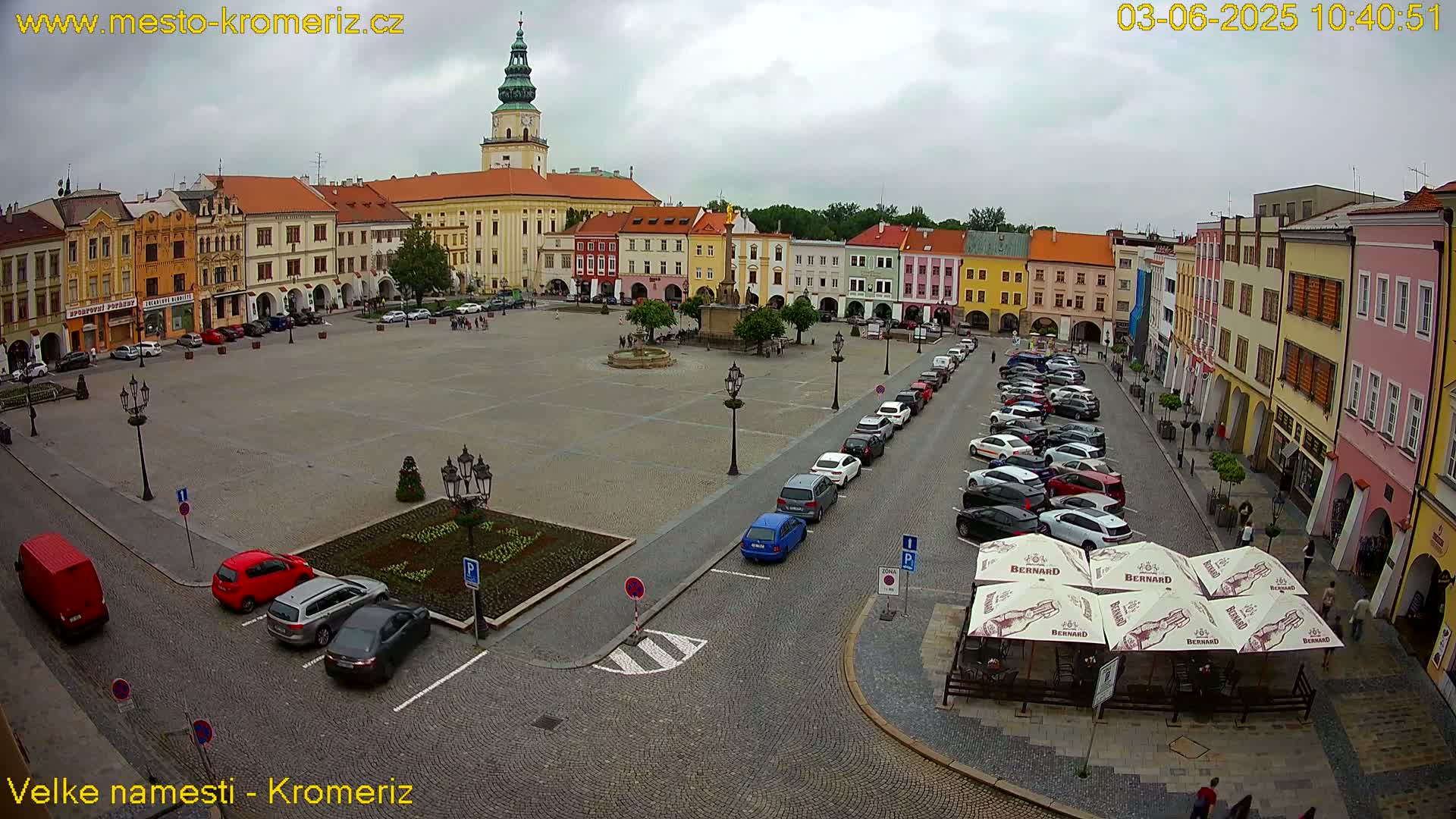 A large town square with colorful buildings under an overcast sky, filled with parked cars and a few pedestrians.