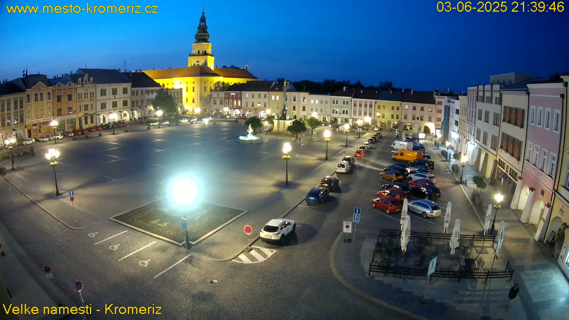 A town square at night, illuminated by streetlights, with cars parked around the perimeter and a fountain in the center.