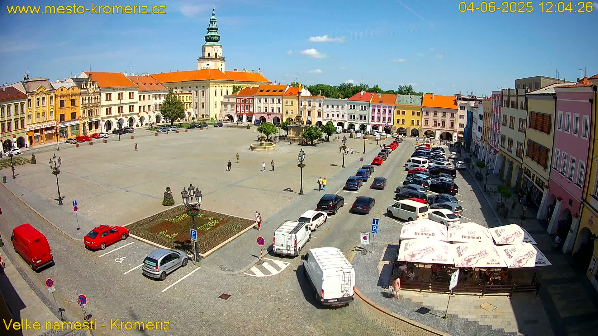 A large town square with colorful buildings surrounding it, filled with parked cars and a few pedestrians, under a bright sunny sky.