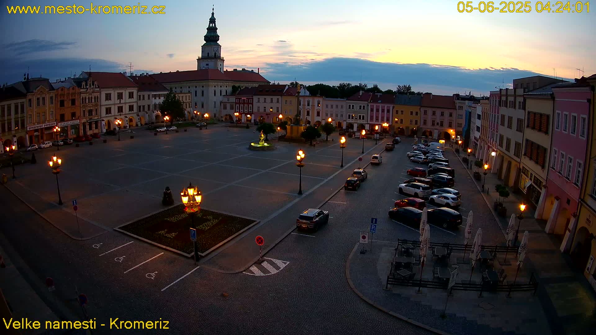 A town square at twilight, illuminated by streetlights, features buildings lining the perimeter and a few cars parked nearby.