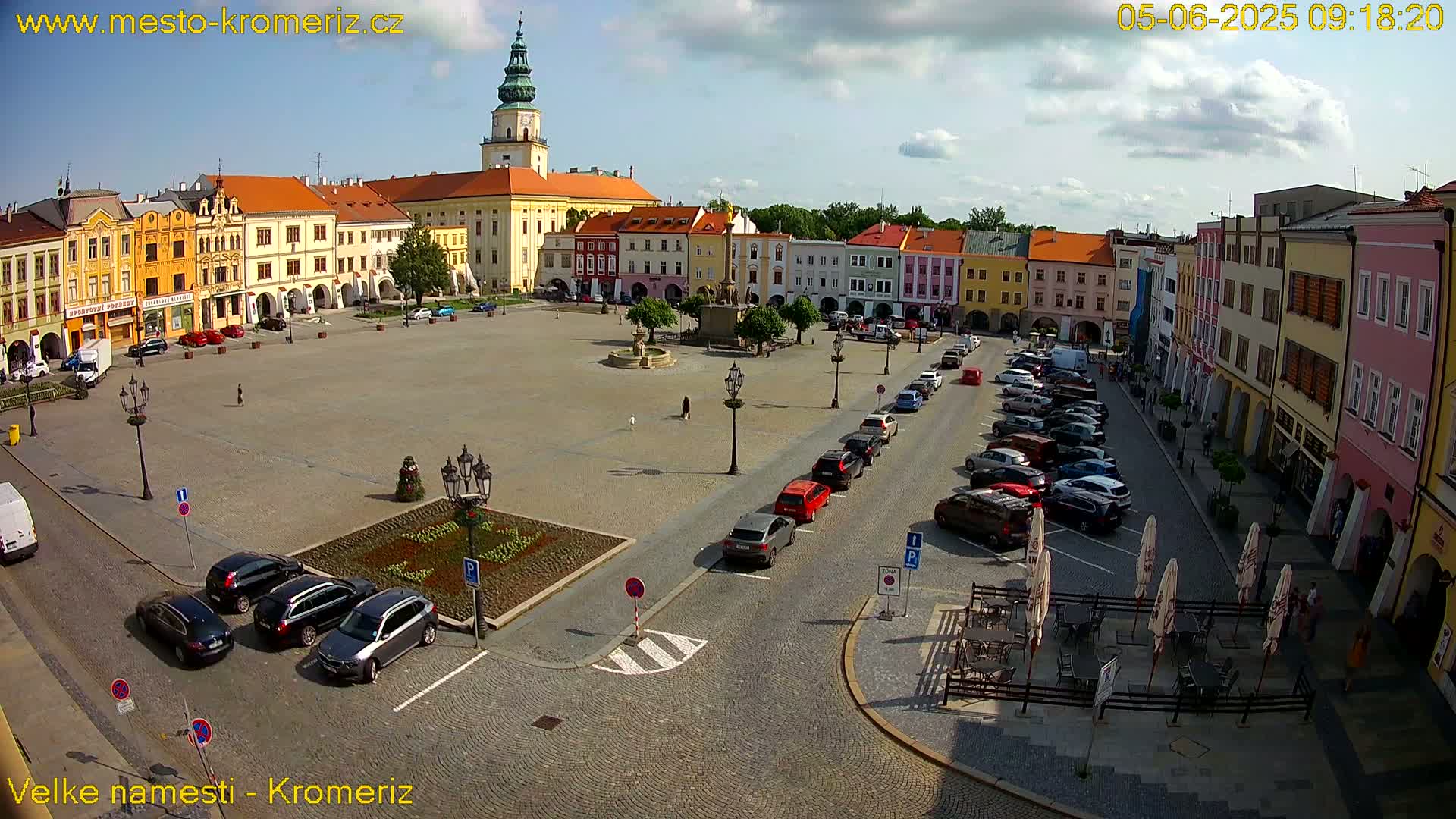 A large town square with colorful buildings surrounding it, a fountain in the center, and cars parked along the sides on a partly sunny day.