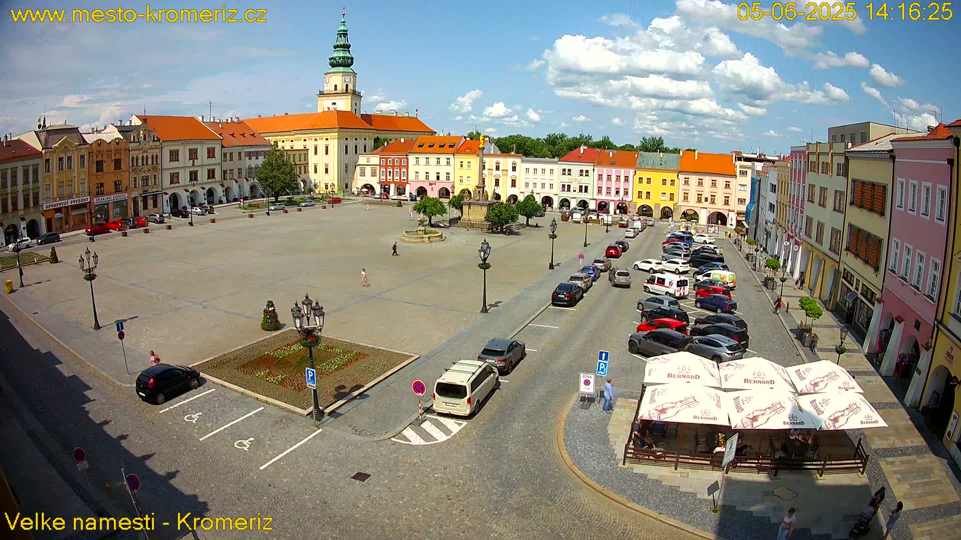 A large town square with colorful buildings surrounding it, filled with parked cars and some pedestrians, under a mostly sunny sky with some clouds.