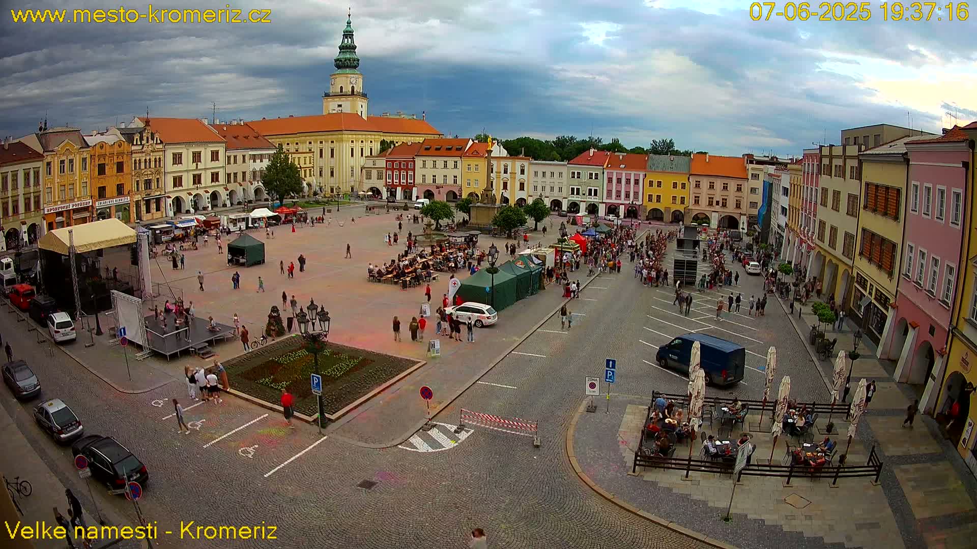 A large town square filled with people, food stalls, and cars under a partly cloudy sky.