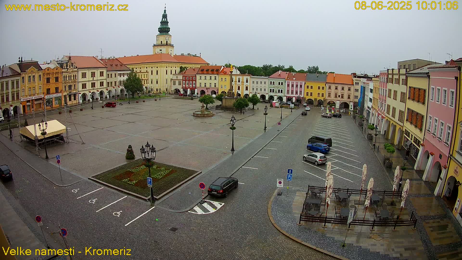 A large town square with colorful buildings surrounding it, a fountain in the center, and several cars parked on the wet cobblestone surface under an overcast sky.