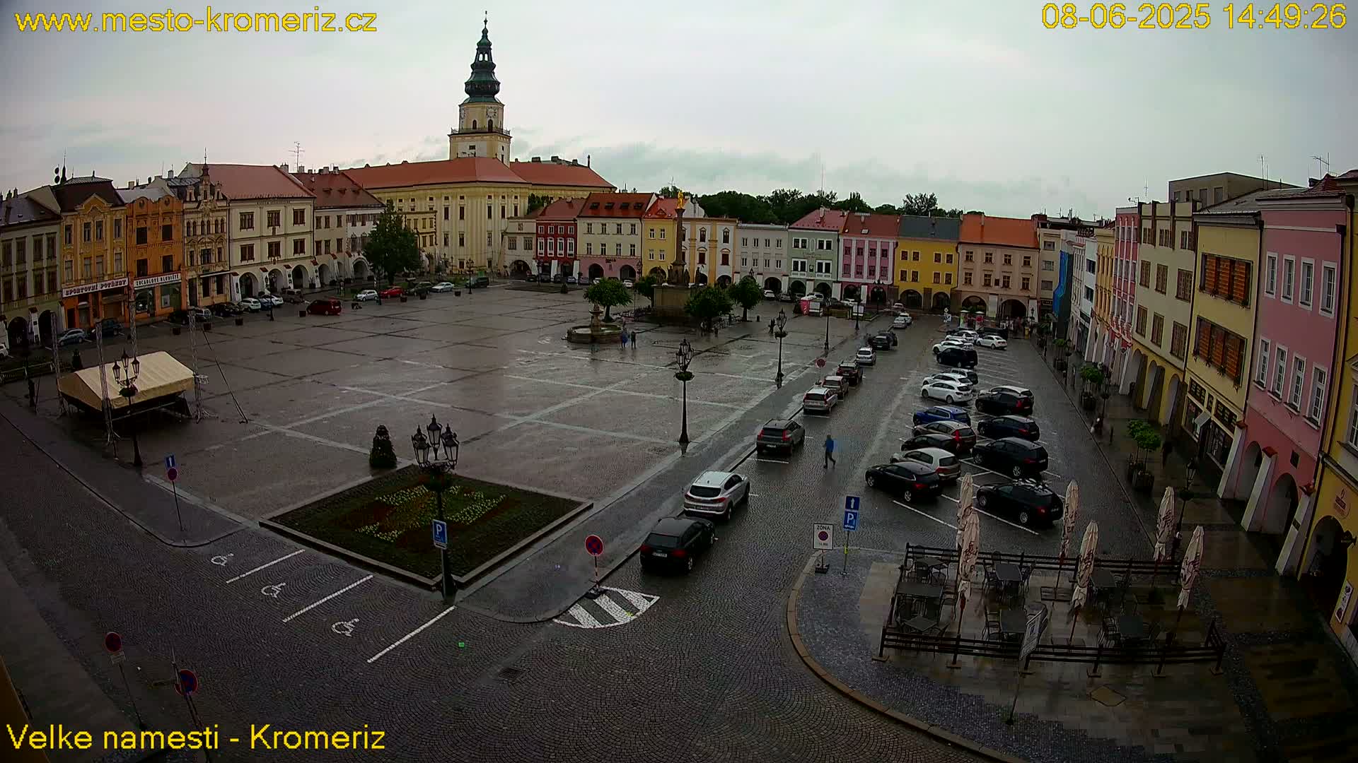 A large town square with colorful buildings surrounding it is wet from a light rain, with cars parked along the sides and a few people walking around.