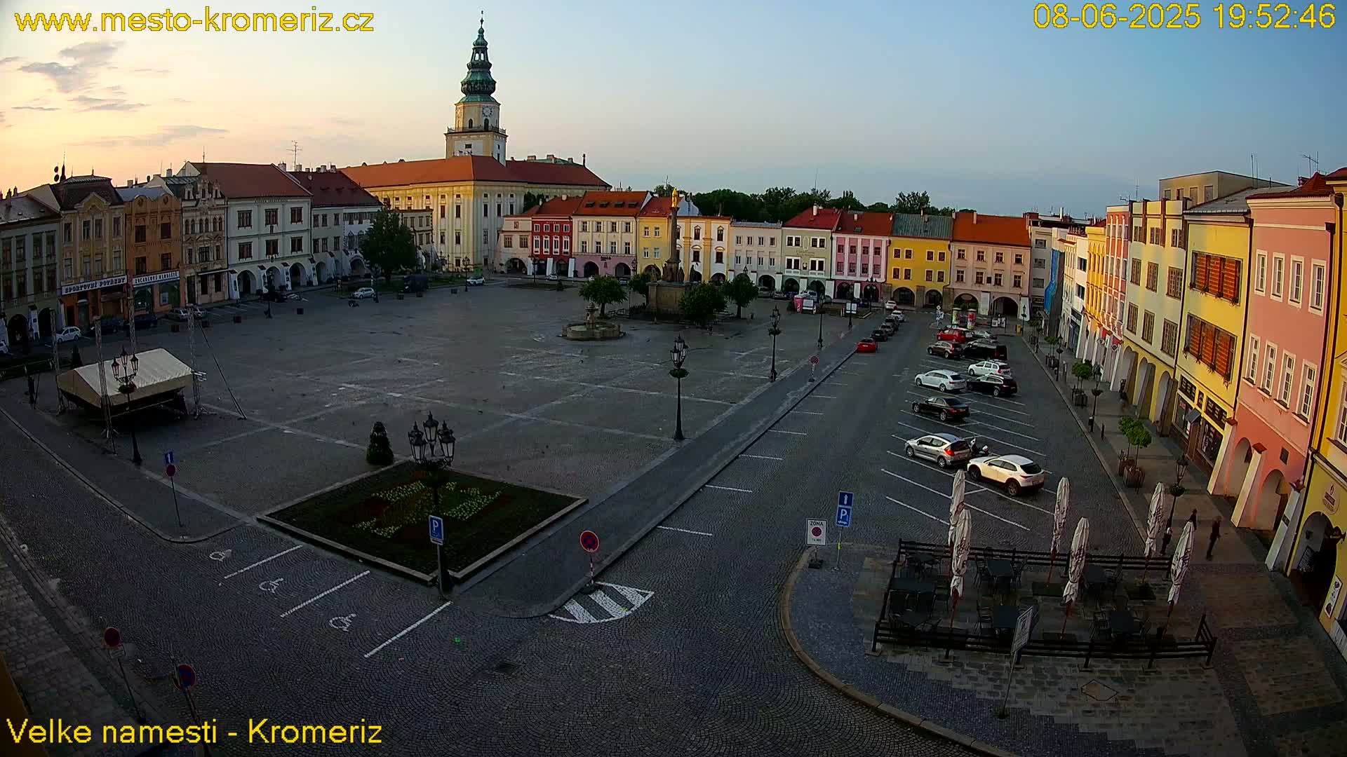 A large town square with colorful buildings surrounding it, a fountain in the center, and cars parked along the edges under a clear, twilight sky.