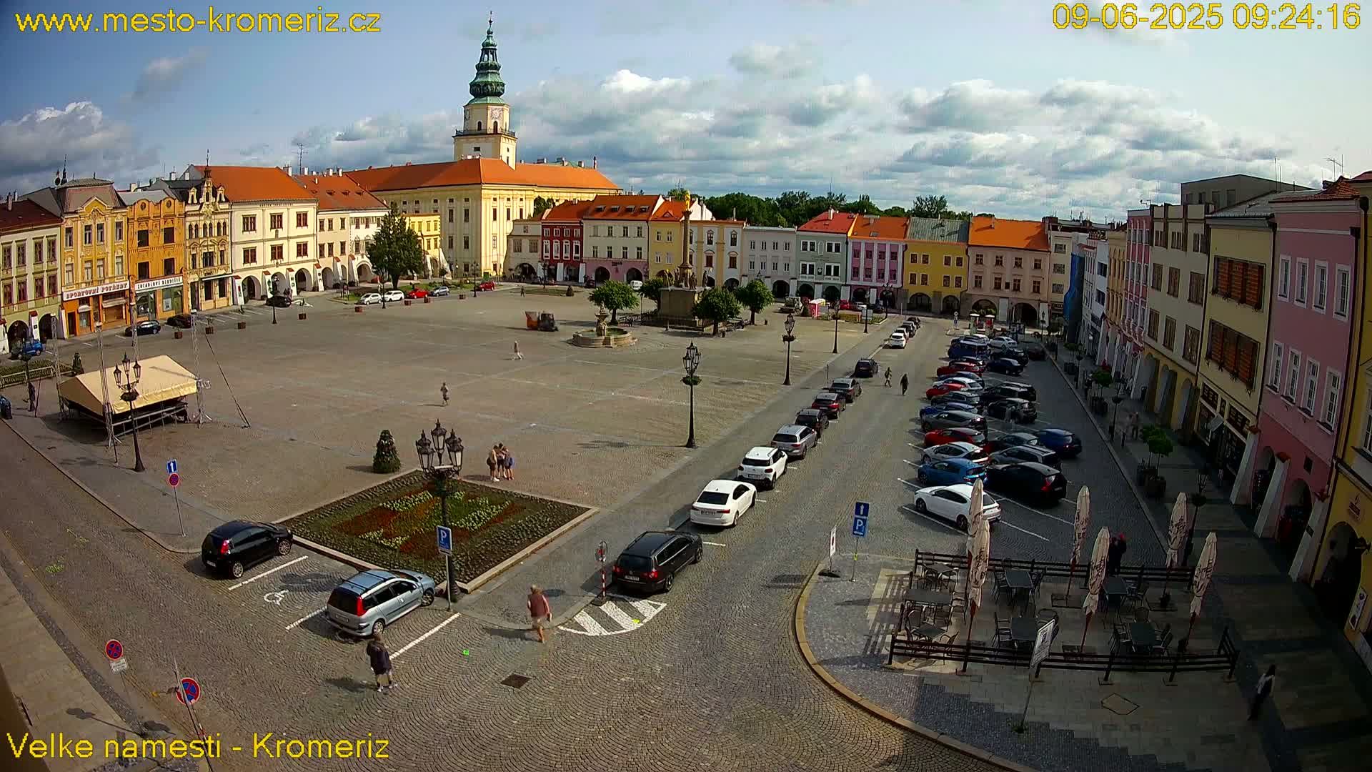 A large town square with colorful buildings surrounding it, a few parked cars, and a partly cloudy sky.