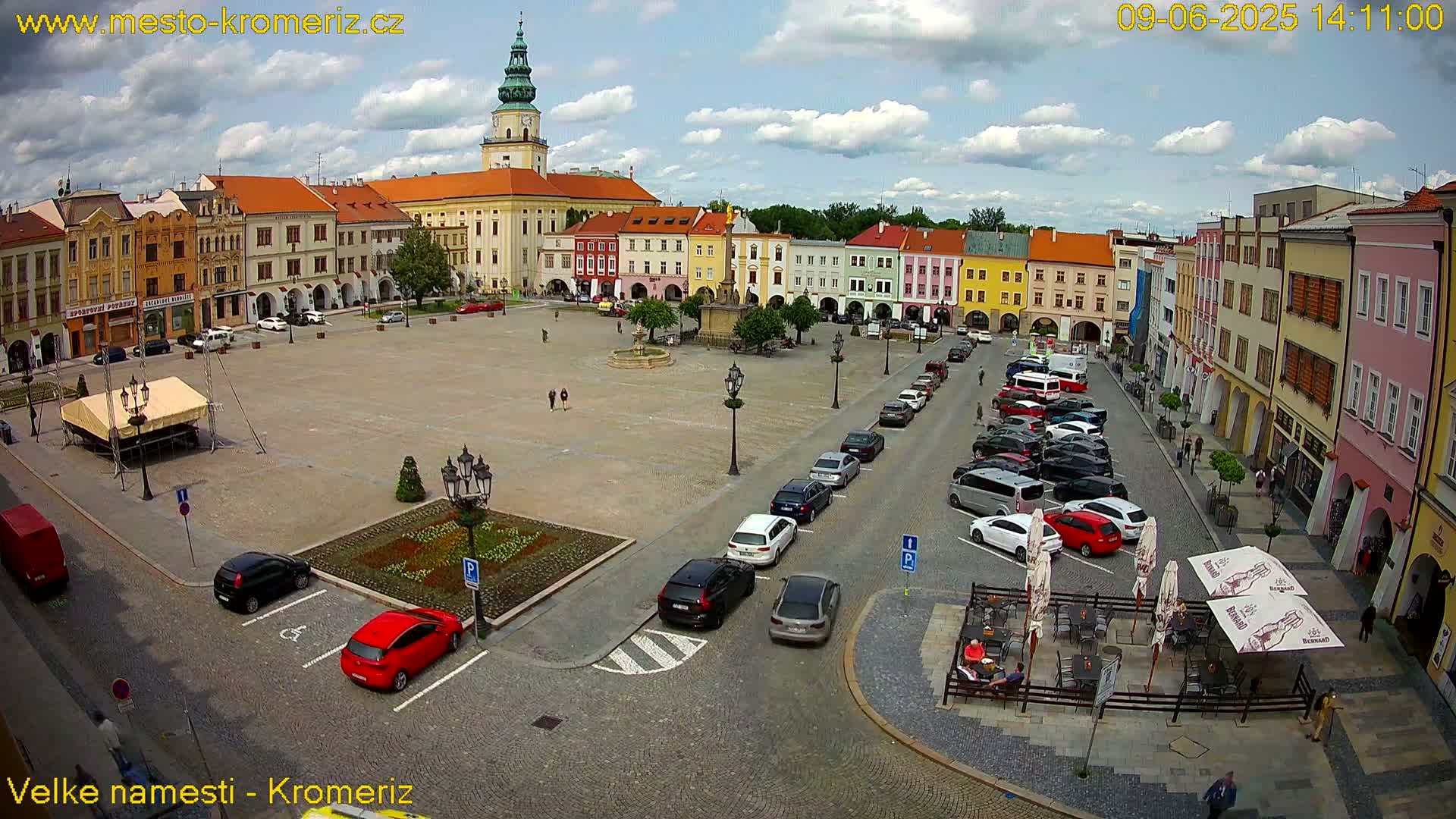 A town square with colorful buildings surrounding it, partly filled with parked cars under a partly cloudy sky.