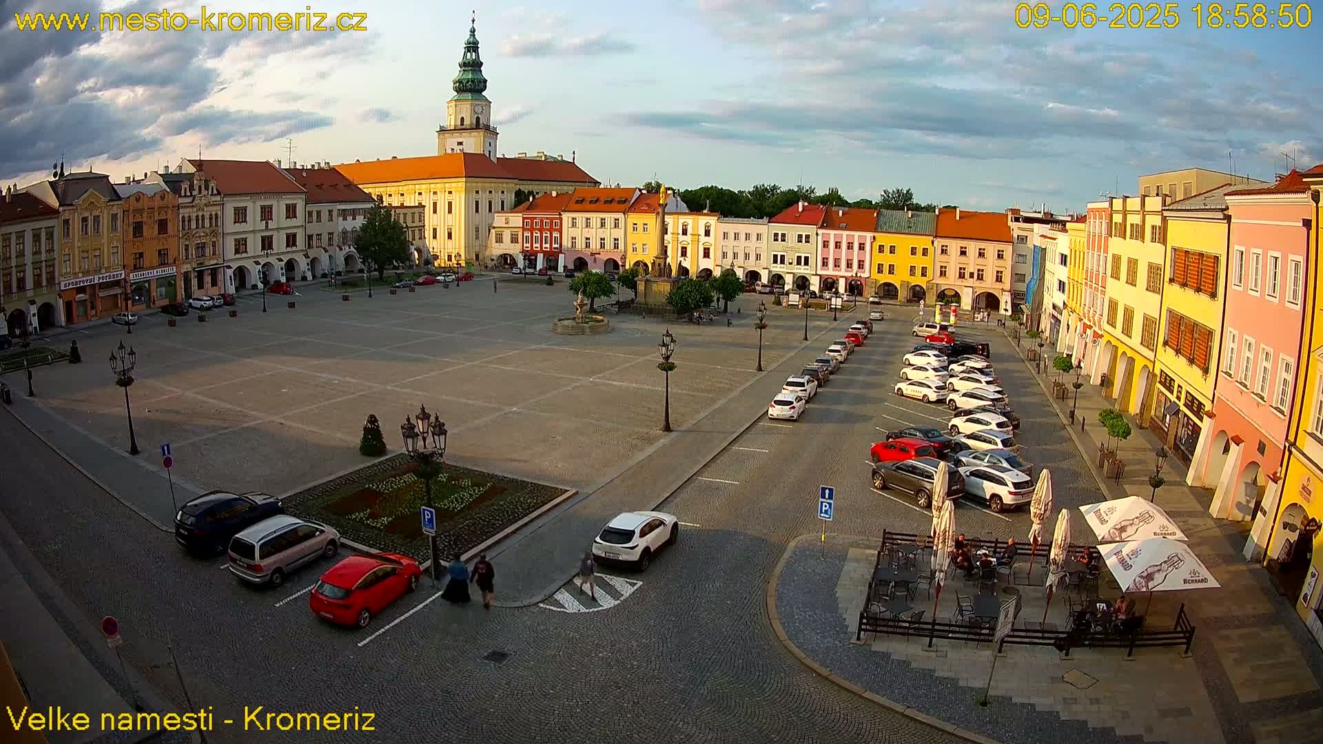 A large town square with colorful buildings, a fountain, parked cars, and an outdoor cafe under a partly cloudy sky.