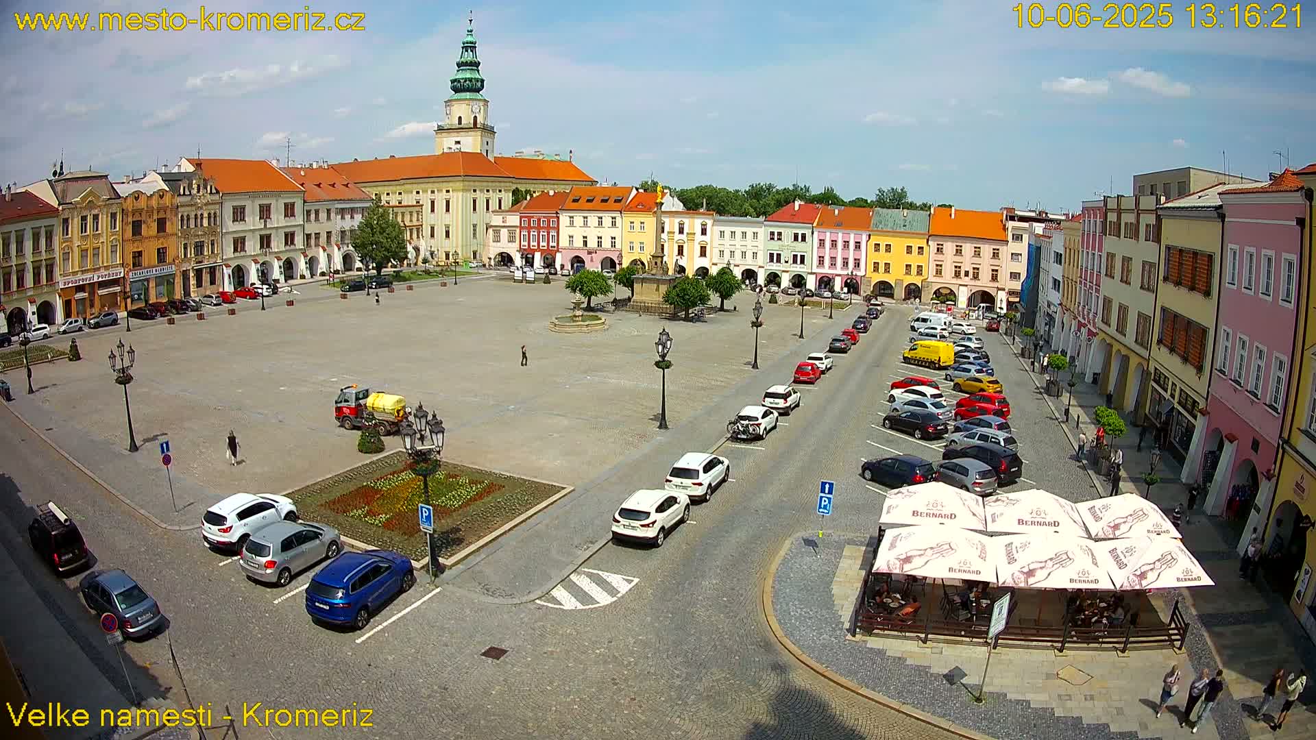 A large town square with colorful buildings surrounding it, several cars parked along the sides, and a small outdoor cafe in the bottom right corner, under a mostly sunny sky.
