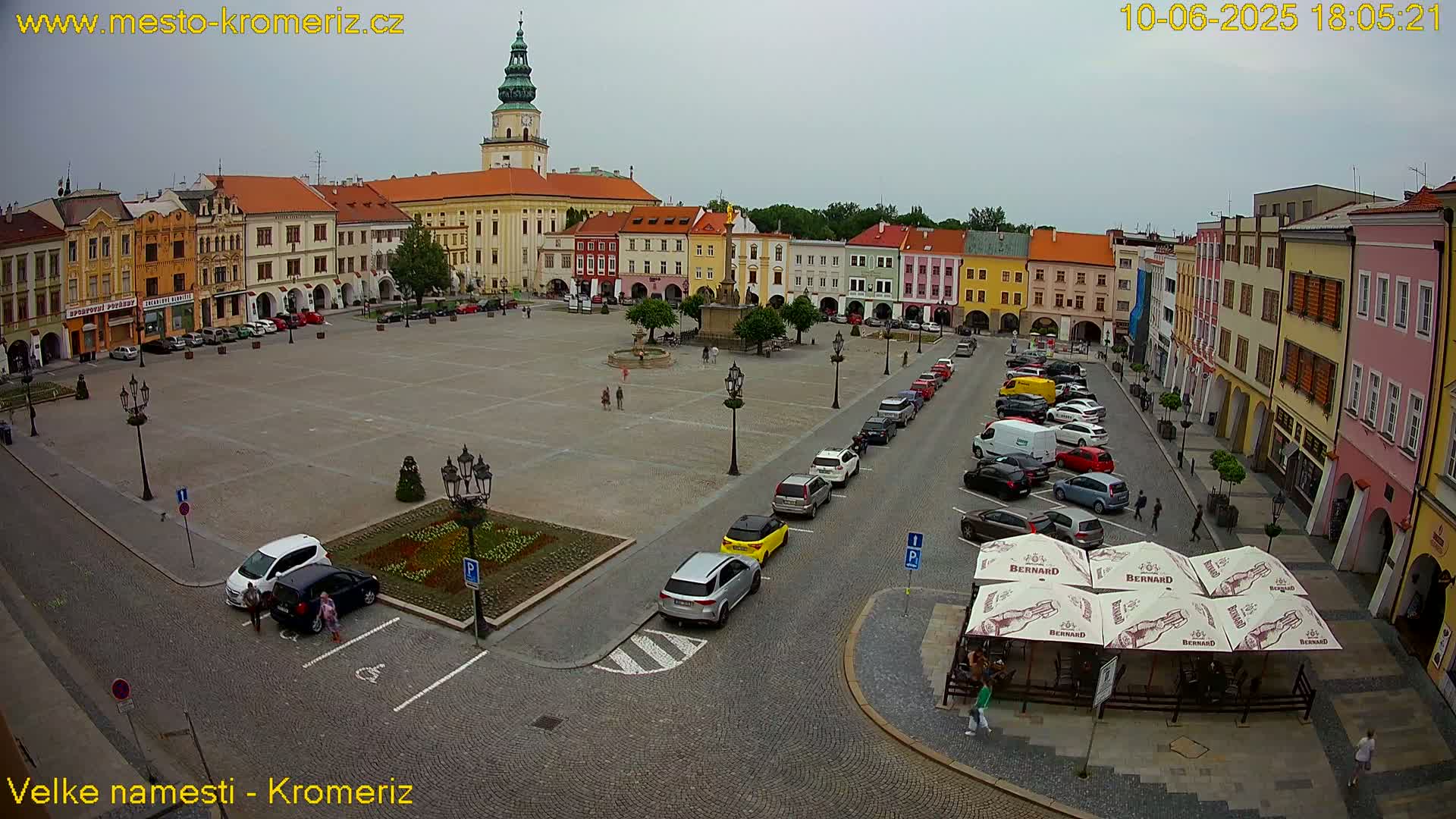 A large town square, surrounded by colorful buildings, under an overcast sky, with cars parked along the sides and a few people walking around.