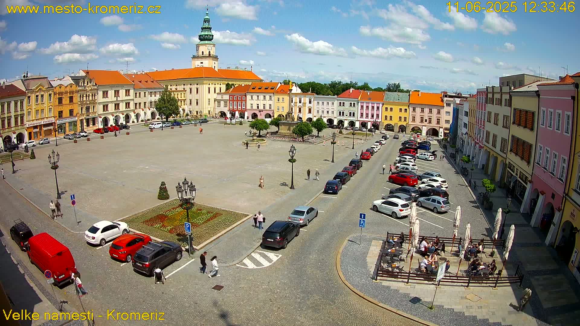A large town square with colorful buildings surrounding it, parked cars, people walking, and outdoor seating at a cafe, under a partly sunny sky.