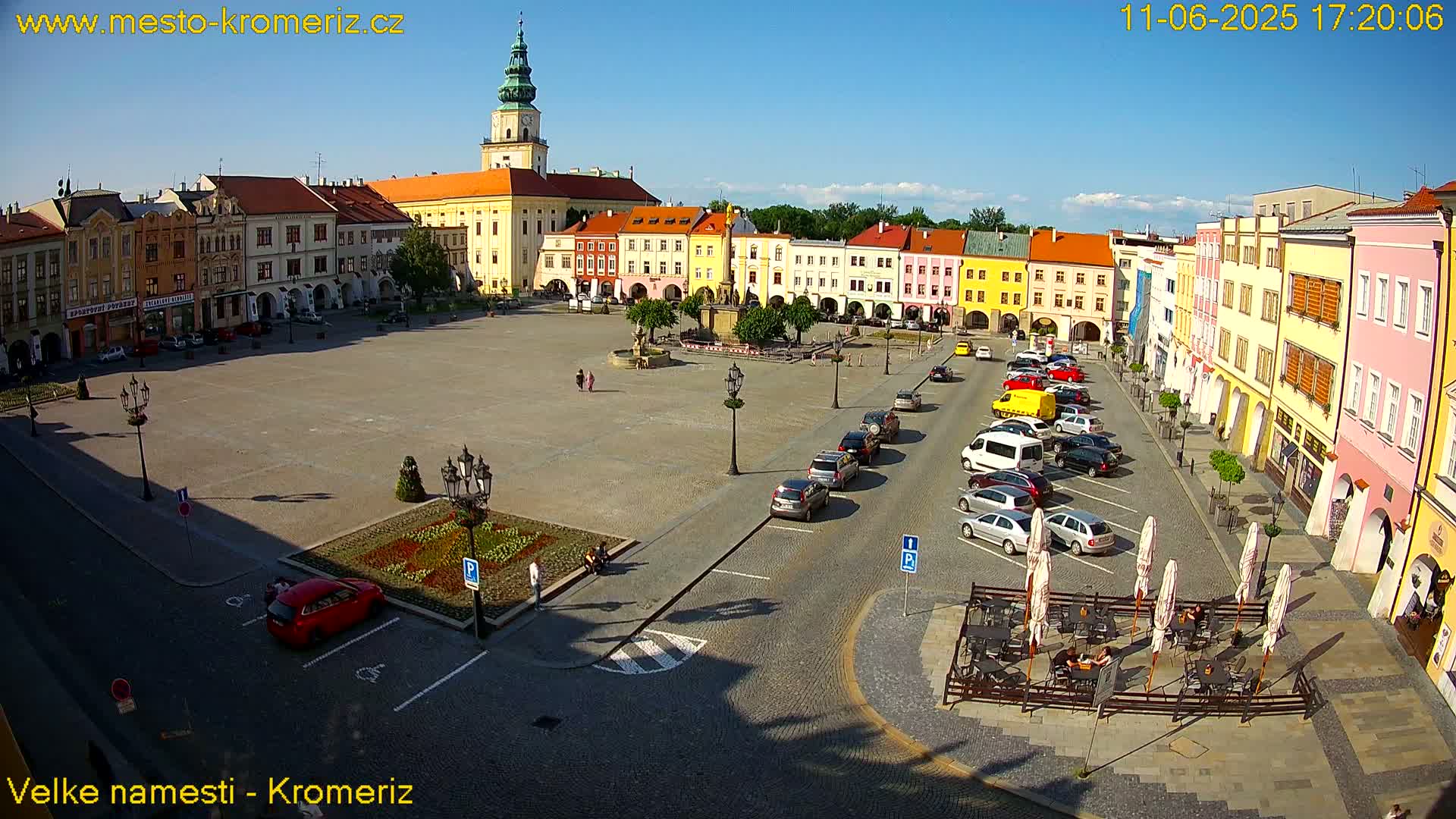 A large town square with colorful buildings surrounding it, cars parked along the sides, and a few people walking around on a sunny day.