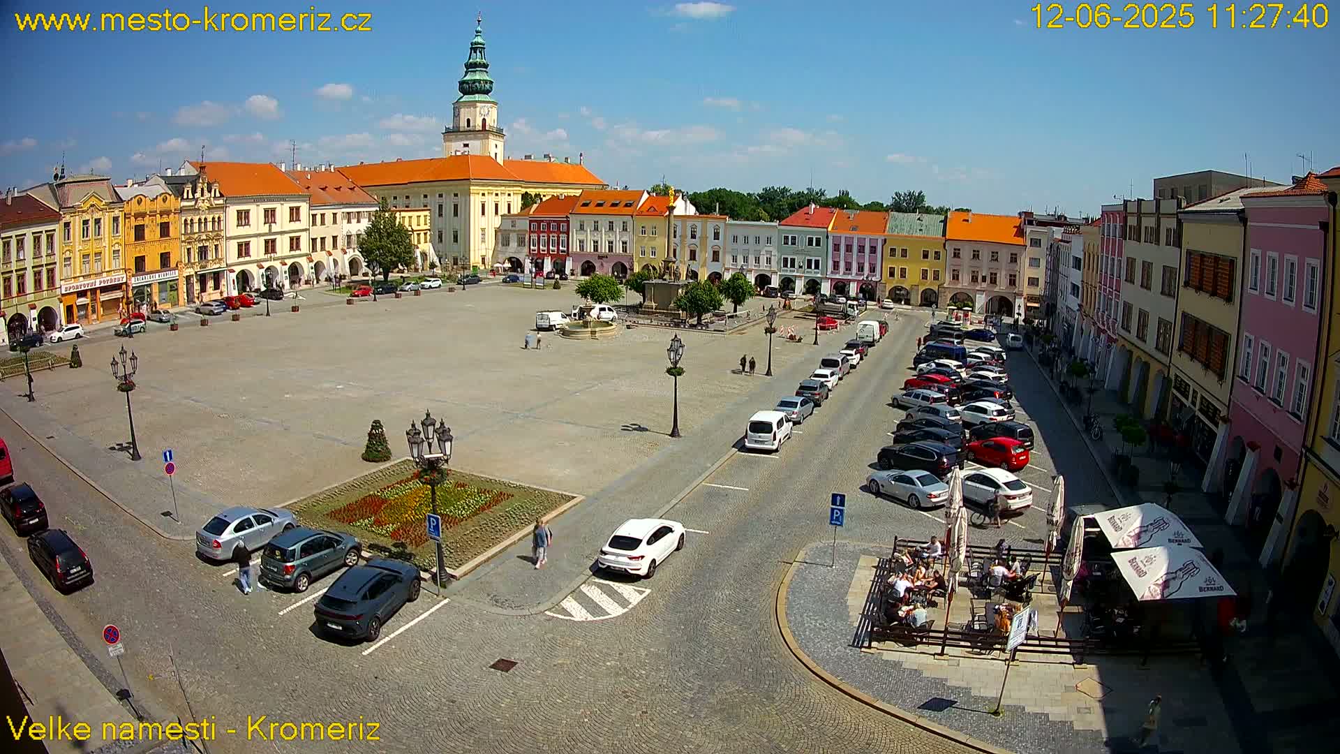 A large town square with colorful buildings surrounding it, a fountain in the center, and cars parked along the sides on a sunny day.