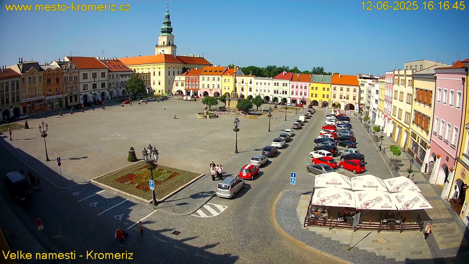 A large town square with colorful buildings surrounding it, under sunny skies, contains parked cars and several people walking around.