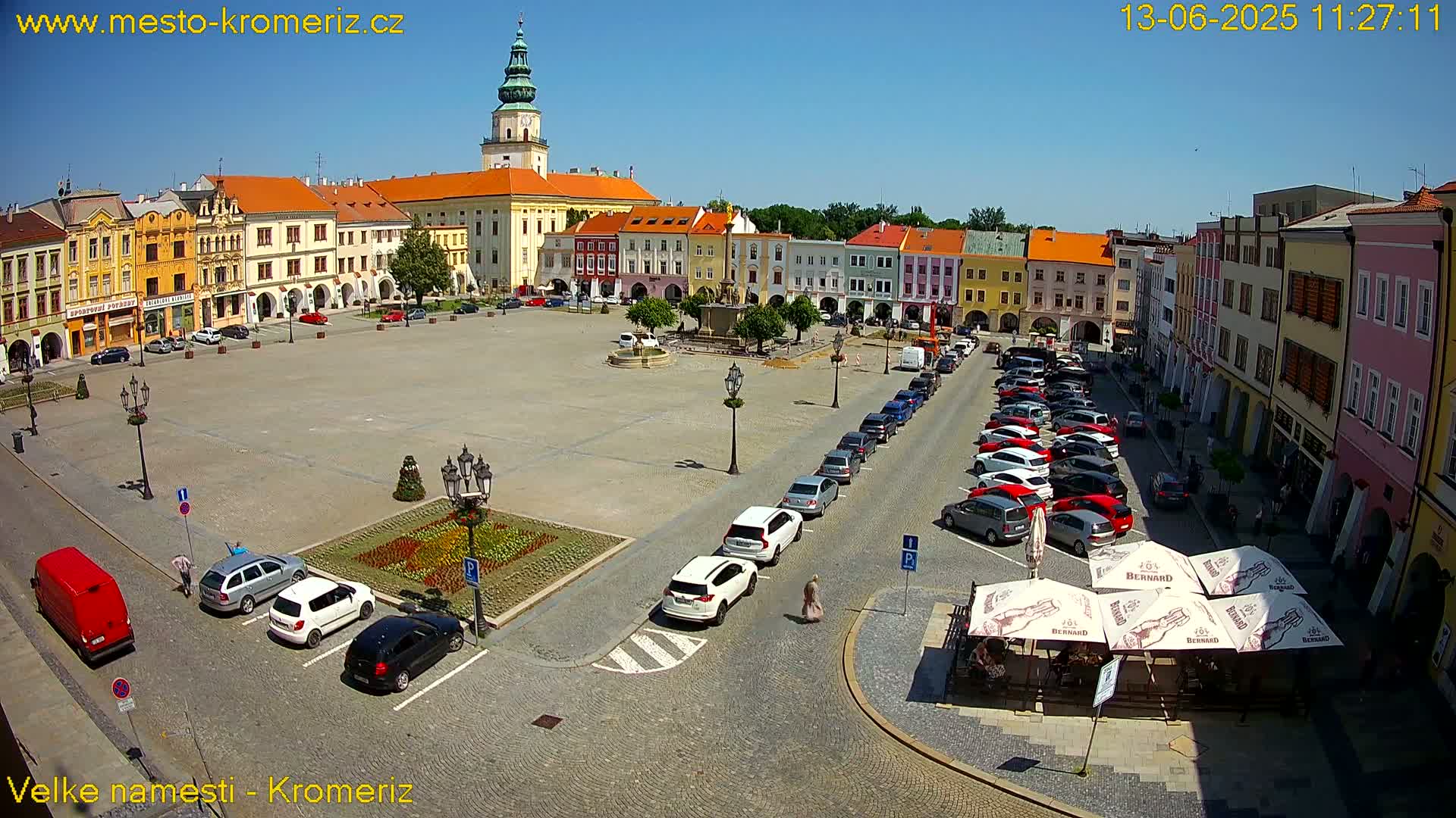 A sunny town square with colorful buildings, a fountain, and parked cars surrounds a large paved area.