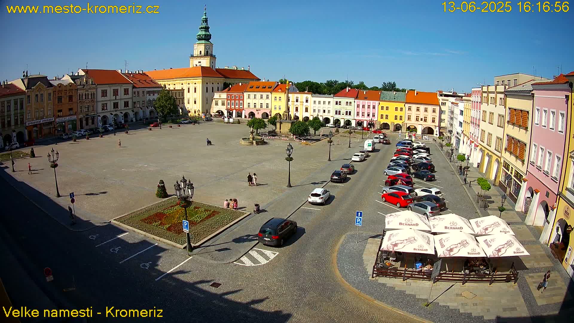 A large town square, filled with parked cars and a few pedestrians, is surrounded by colorful buildings under a clear, sunny sky.