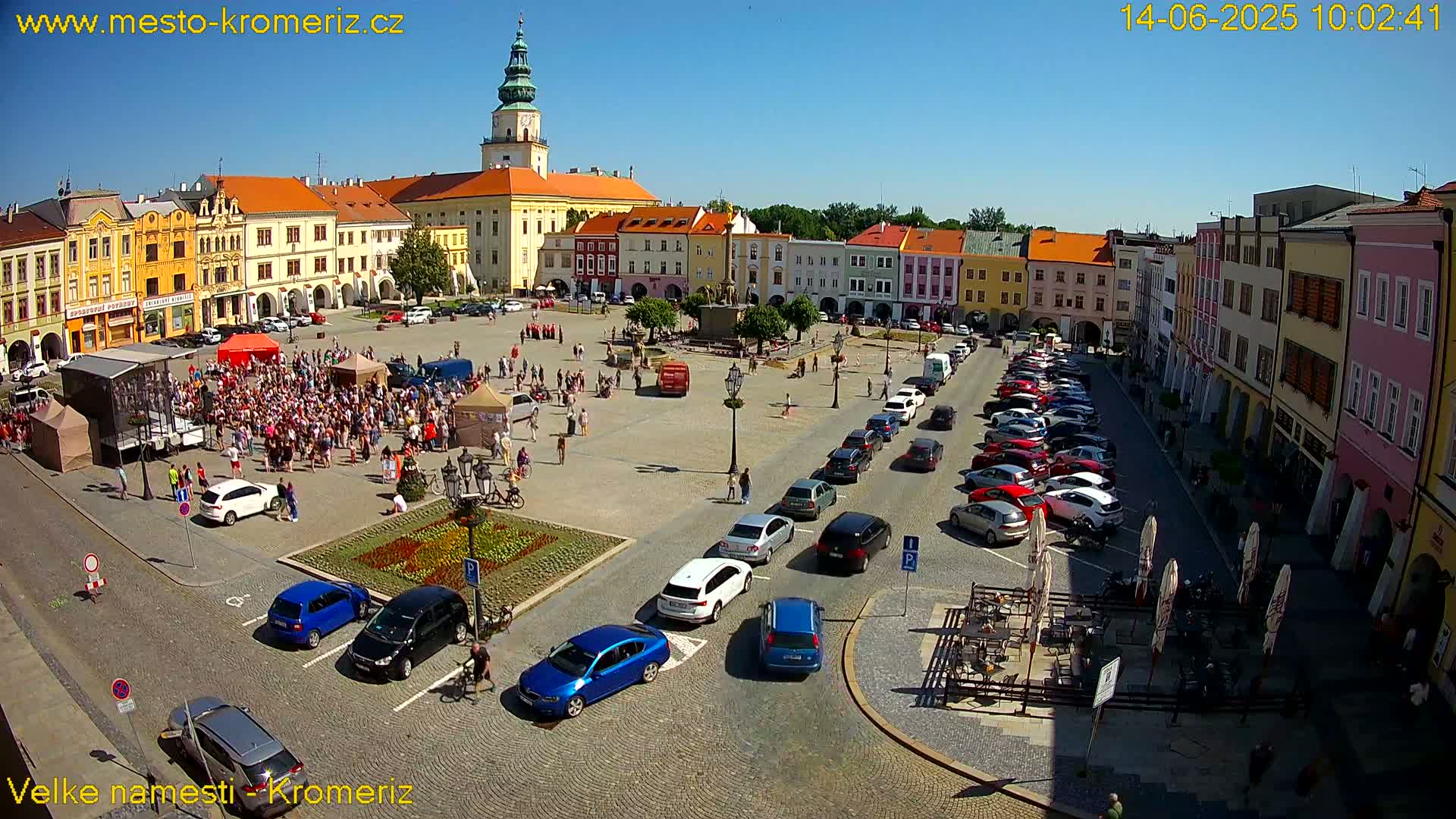 A large town square filled with people, cars parked along the sides, and colorful buildings surrounding it under sunny skies.