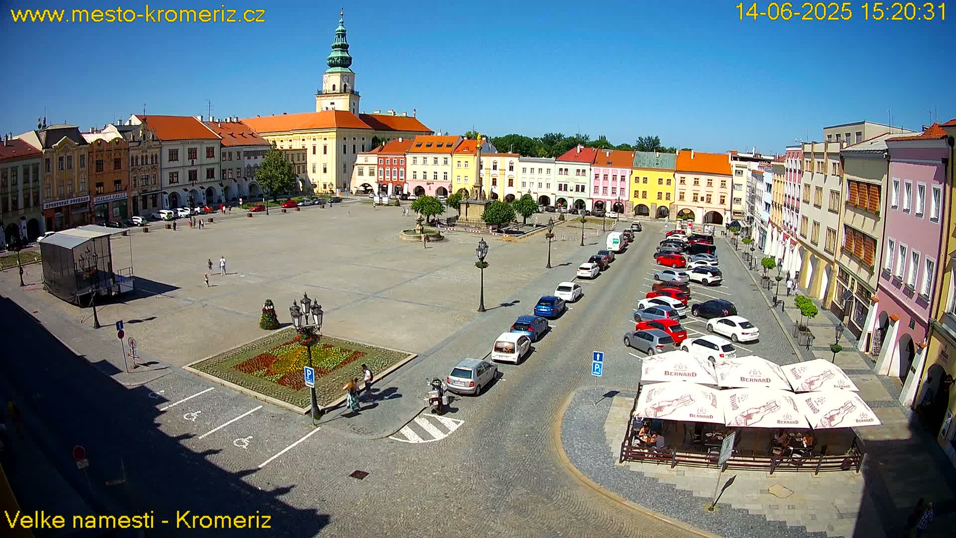 A sunny town square, surrounded by colorful buildings, with parked cars and a few pedestrians.
