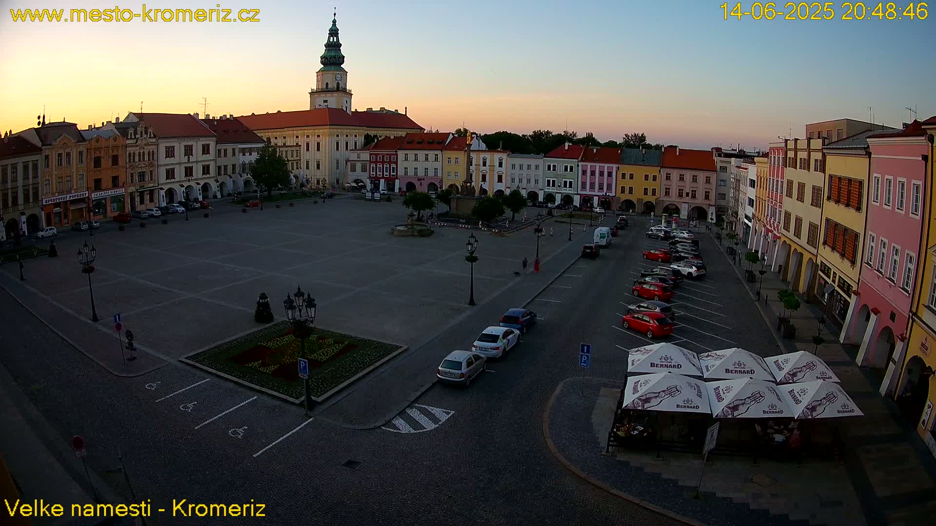 A sunny town square, surrounded by colorful buildings, with parked cars and a few pedestrians.