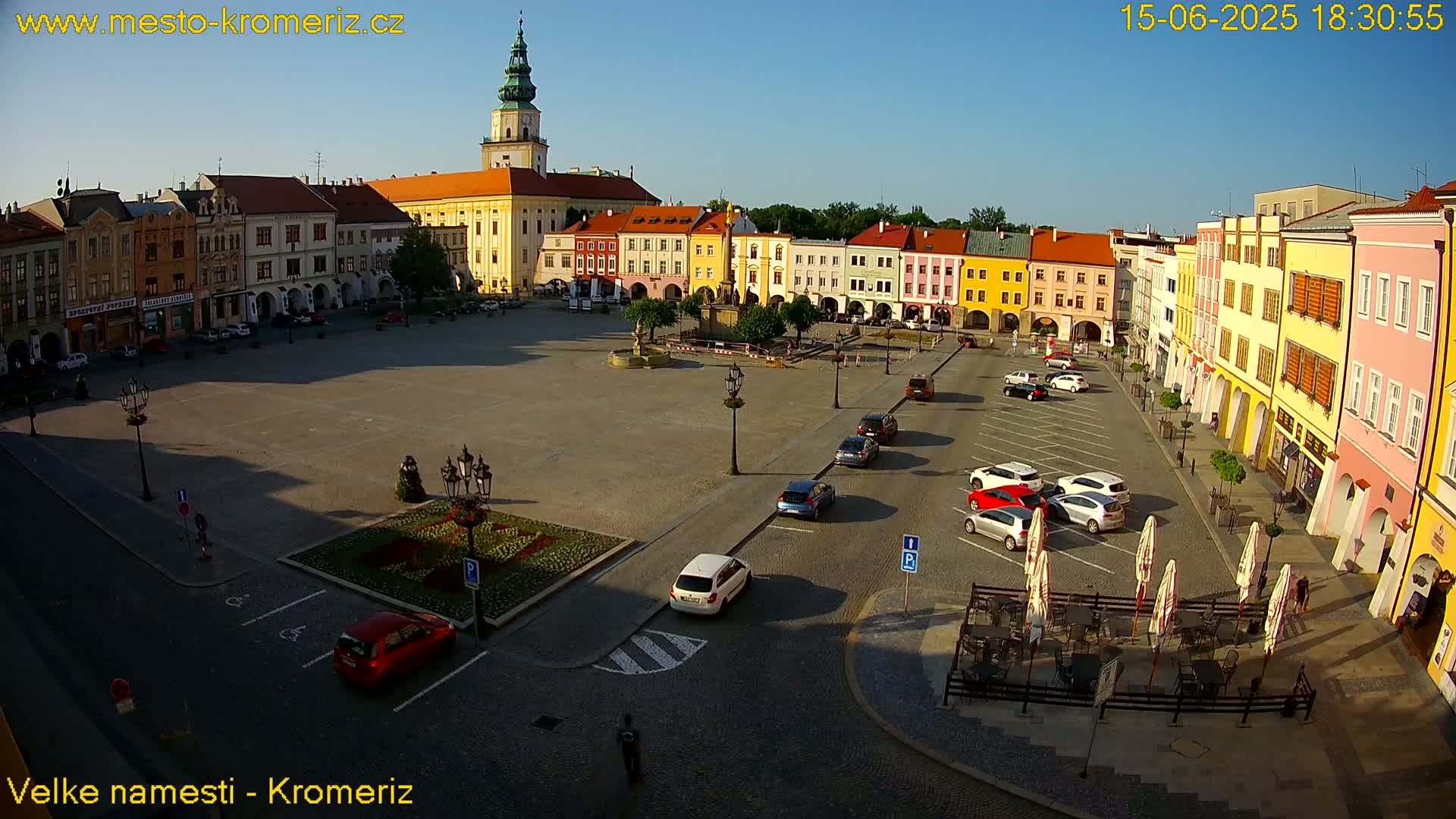 A large town square with colorful buildings surrounding it, a few cars parked, and outdoor seating at a cafe, all under a sunny sky.