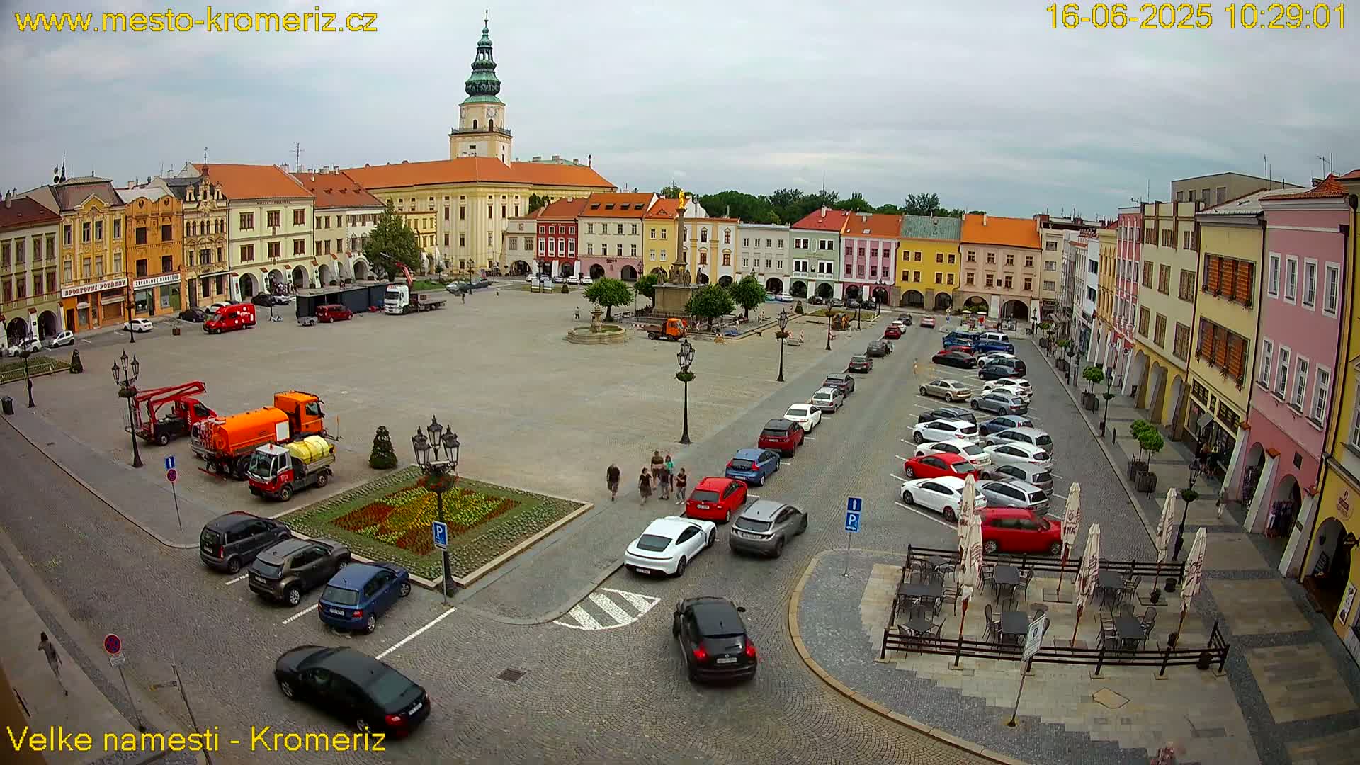 A large town square with colorful buildings surrounding it, under an overcast sky, shows several cars parked and a few people walking, along with some street cleaning vehicles.