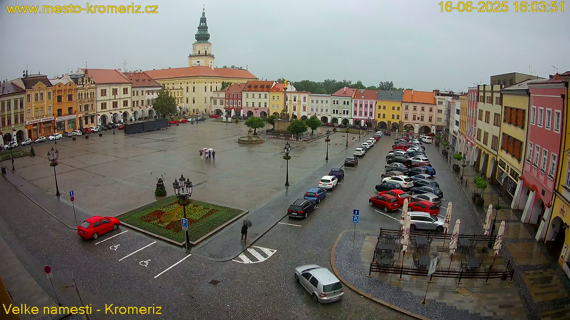 A rain-soaked town square, surrounded by colorful buildings and parked cars, features a central fountain and several people with umbrellas.