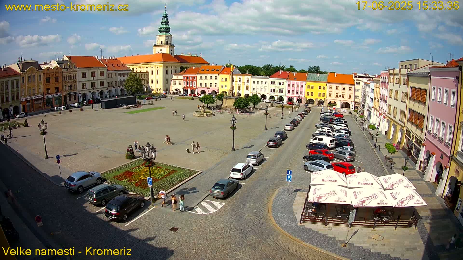 A large town square with colorful buildings surrounding it, many cars parked along the sides, and a few people walking around on a sunny day.