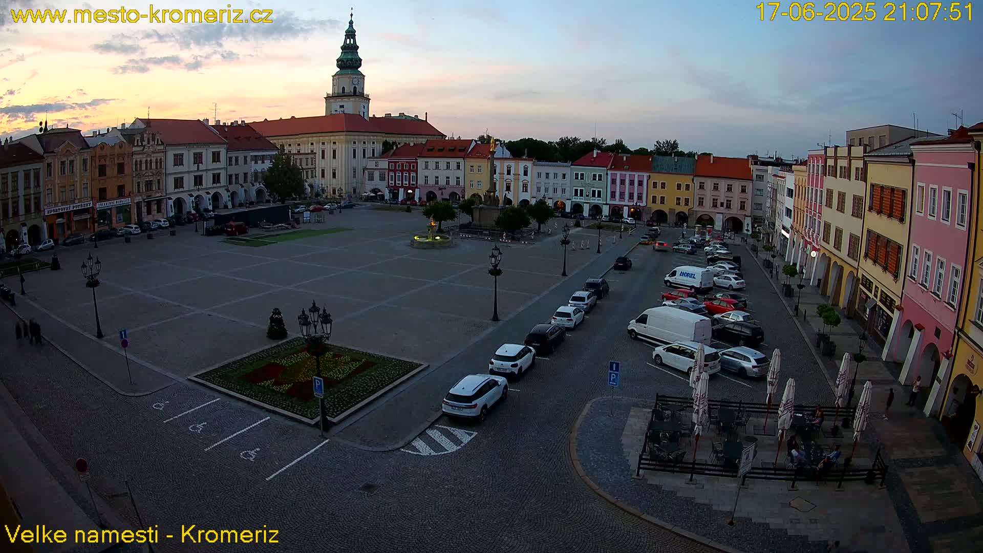 A large town square with colorful buildings surrounding it, cars parked along the sides, and a few people sitting at outdoor tables under parasols, during a twilight sky.