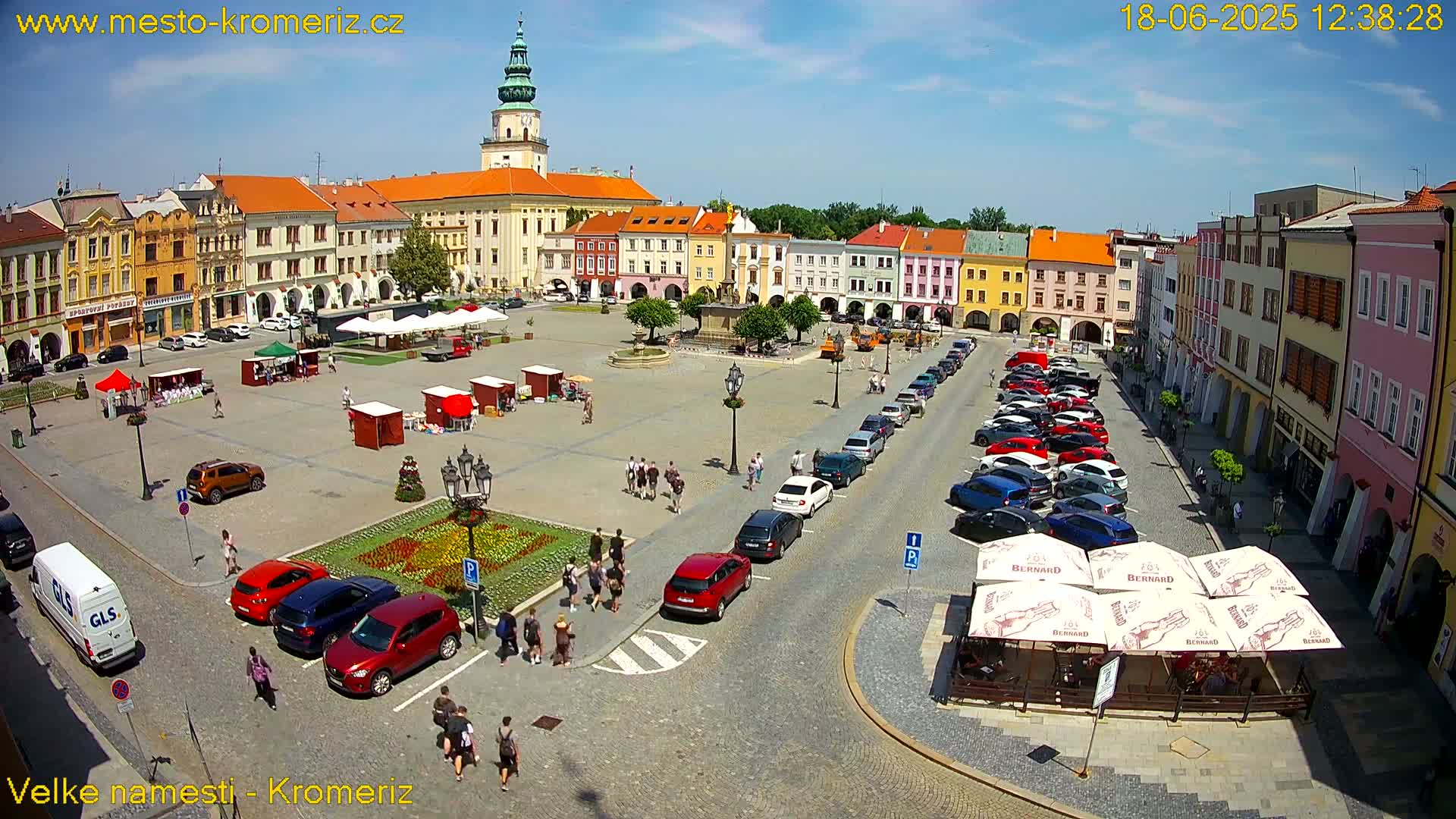 A sunny town square with colorful buildings, parked cars, and several market stalls, surrounded by pedestrians.