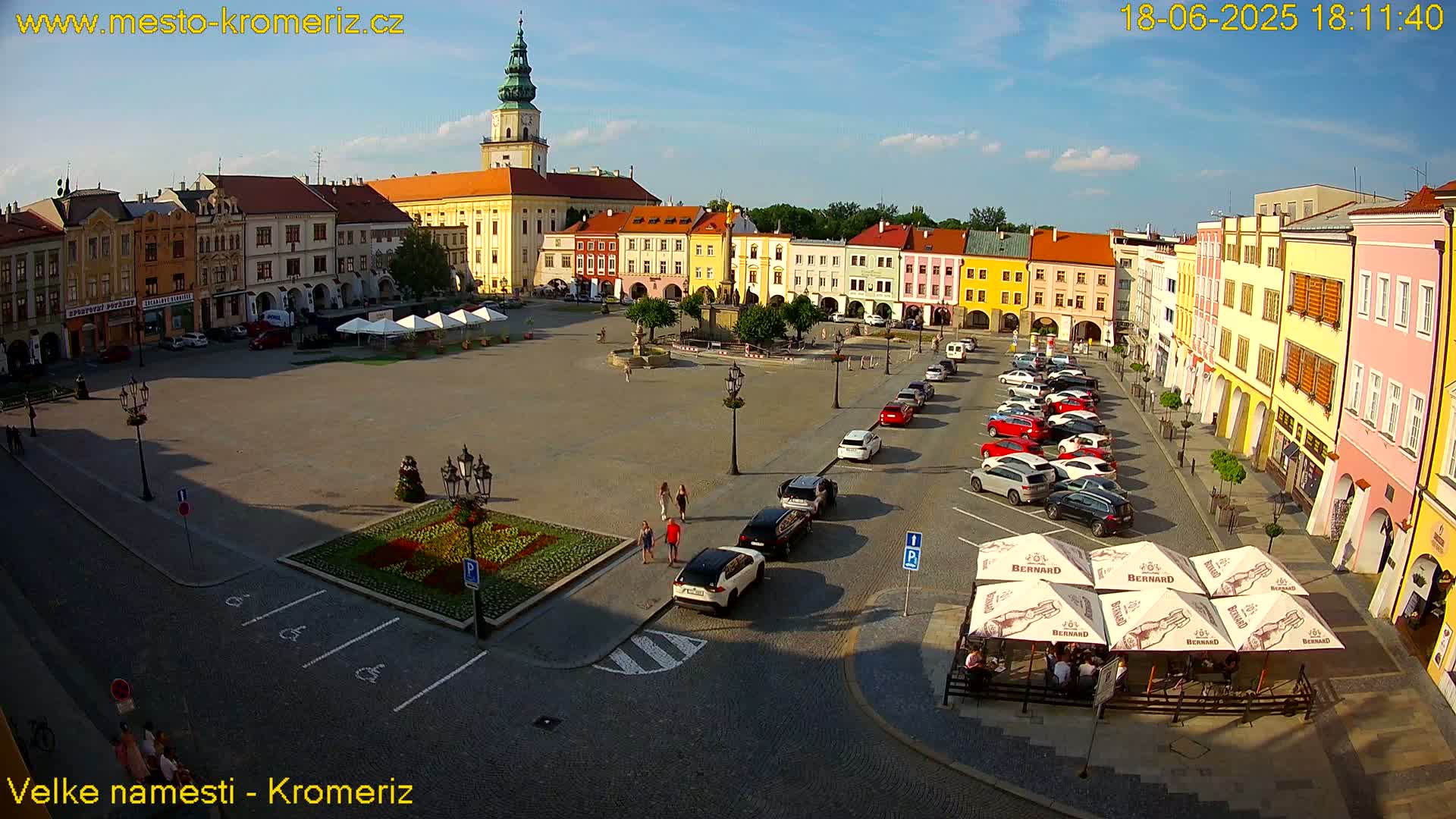 A town square, surrounded by colorful buildings under a mostly sunny sky, features parked cars and several people walking around.