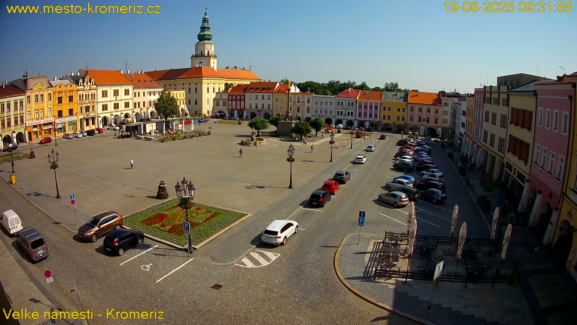 A large town square with colorful buildings surrounding it, filled with parked cars and a few pedestrians, under a clear sunny sky.