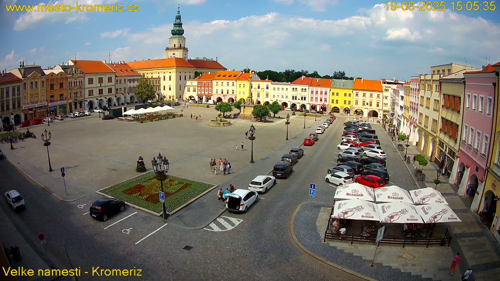 A large town square with colorful buildings surrounding it, many cars parked along the sides, and a sunny day.
