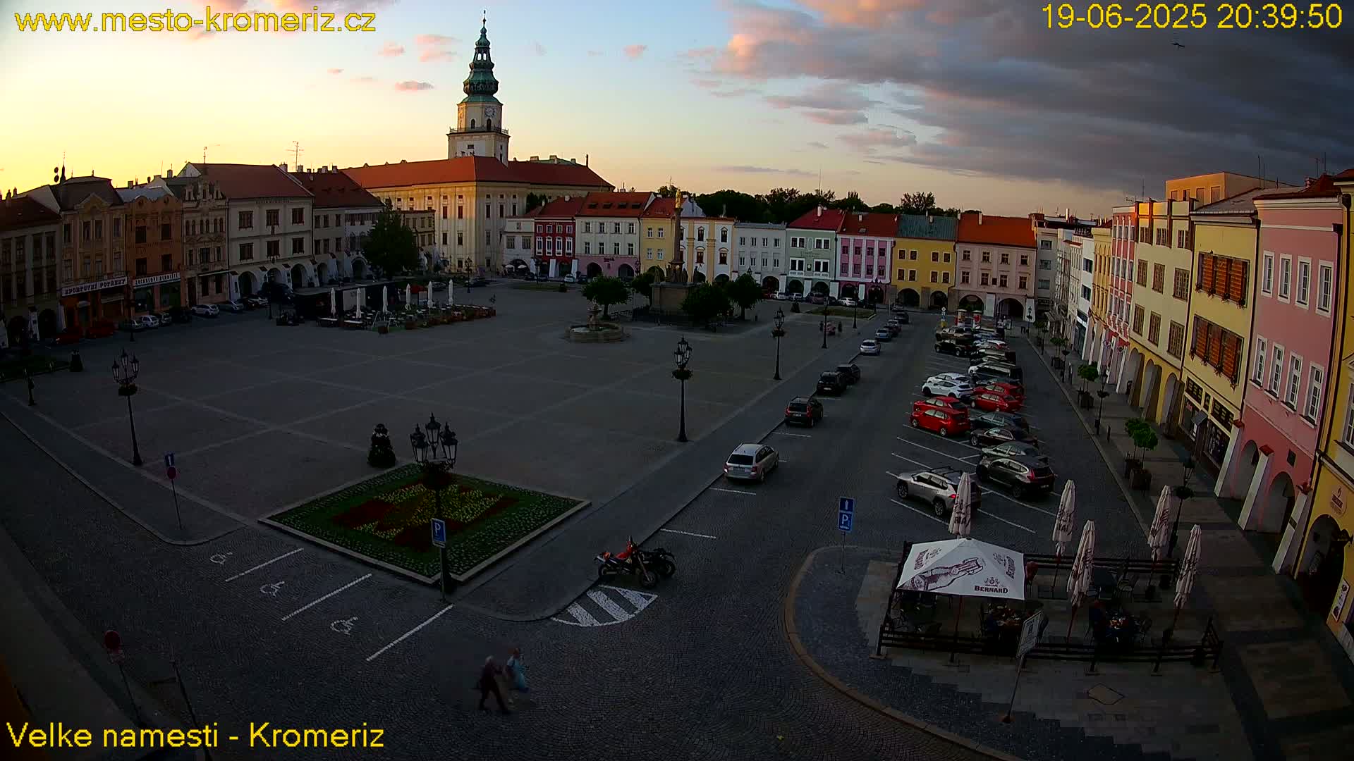 A large town square with colorful buildings surrounding it, cars parked along the sides, and a few people walking around under a partly cloudy sunset sky.