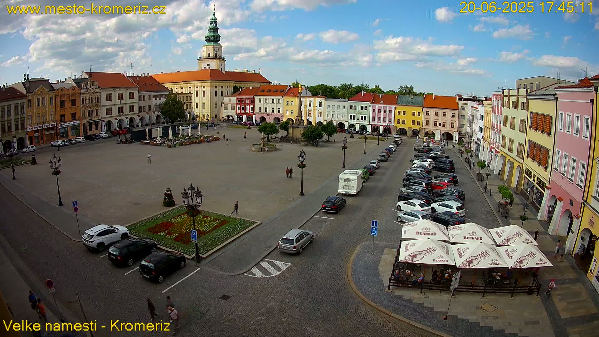 A large town square with colorful buildings surrounding it, under a mostly sunny sky with some clouds, has cars parked along the sides and people walking around.