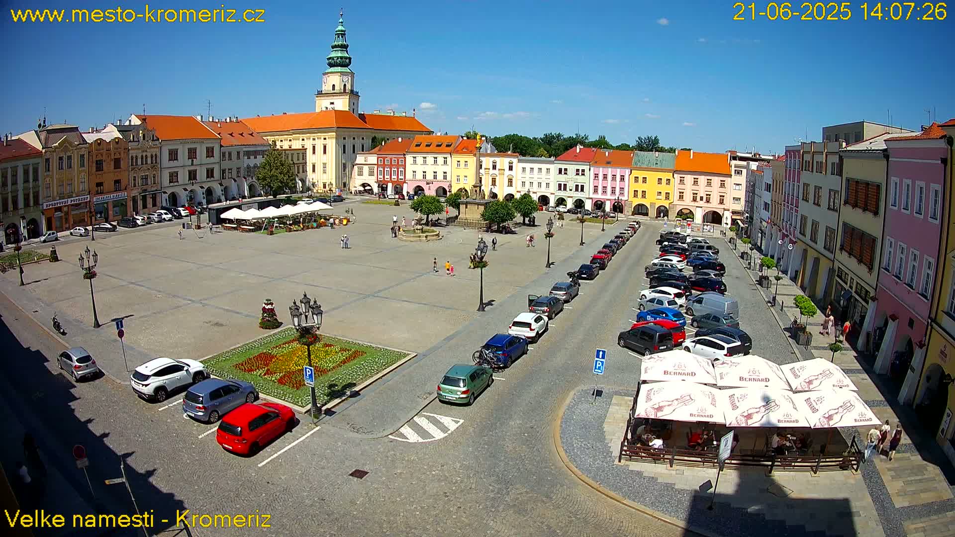 A large town square with colorful buildings, a fountain, parked cars, and outdoor seating under umbrellas on a sunny day.