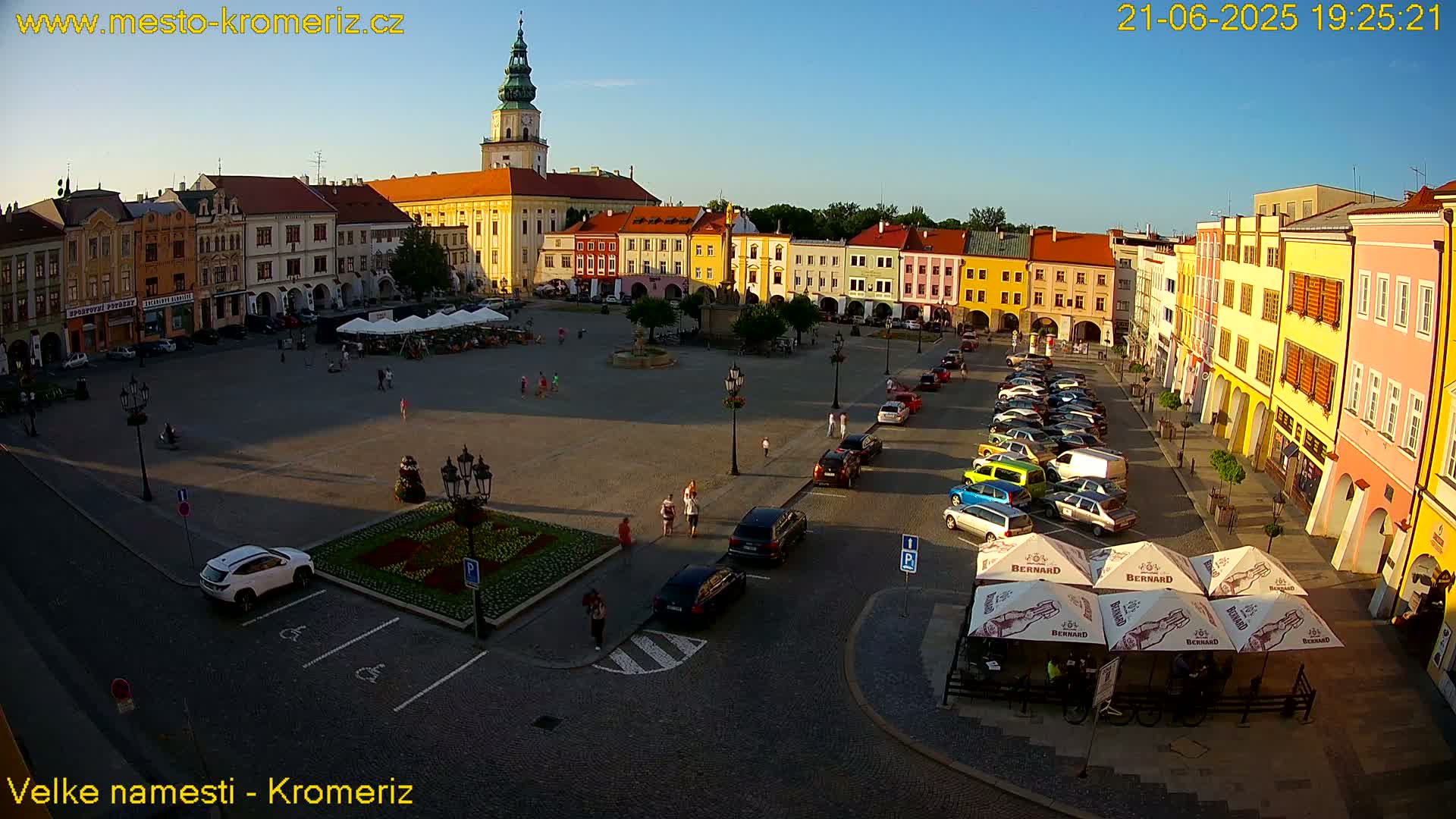 A large town square, surrounded by colorful buildings, is filled with parked cars and a few pedestrians under a clear, sunny sky.
