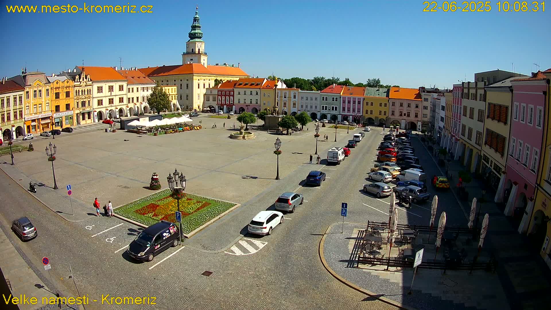 A large town square with colorful buildings surrounding it, cars parked along the sides, and a few people walking around on a sunny day.