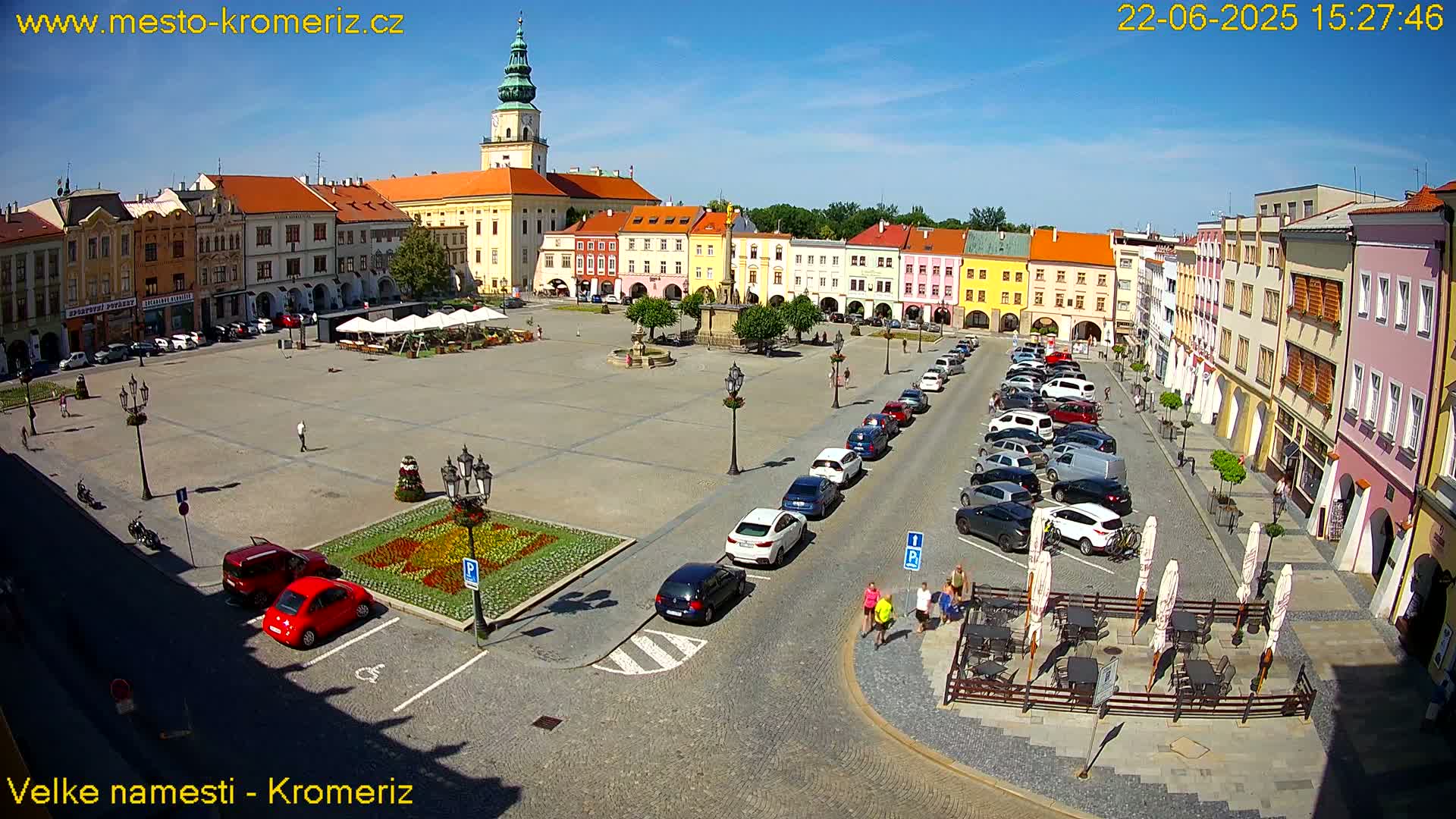 A town square with colorful buildings, a fountain, parked cars, and outdoor seating under umbrellas on a sunny day.