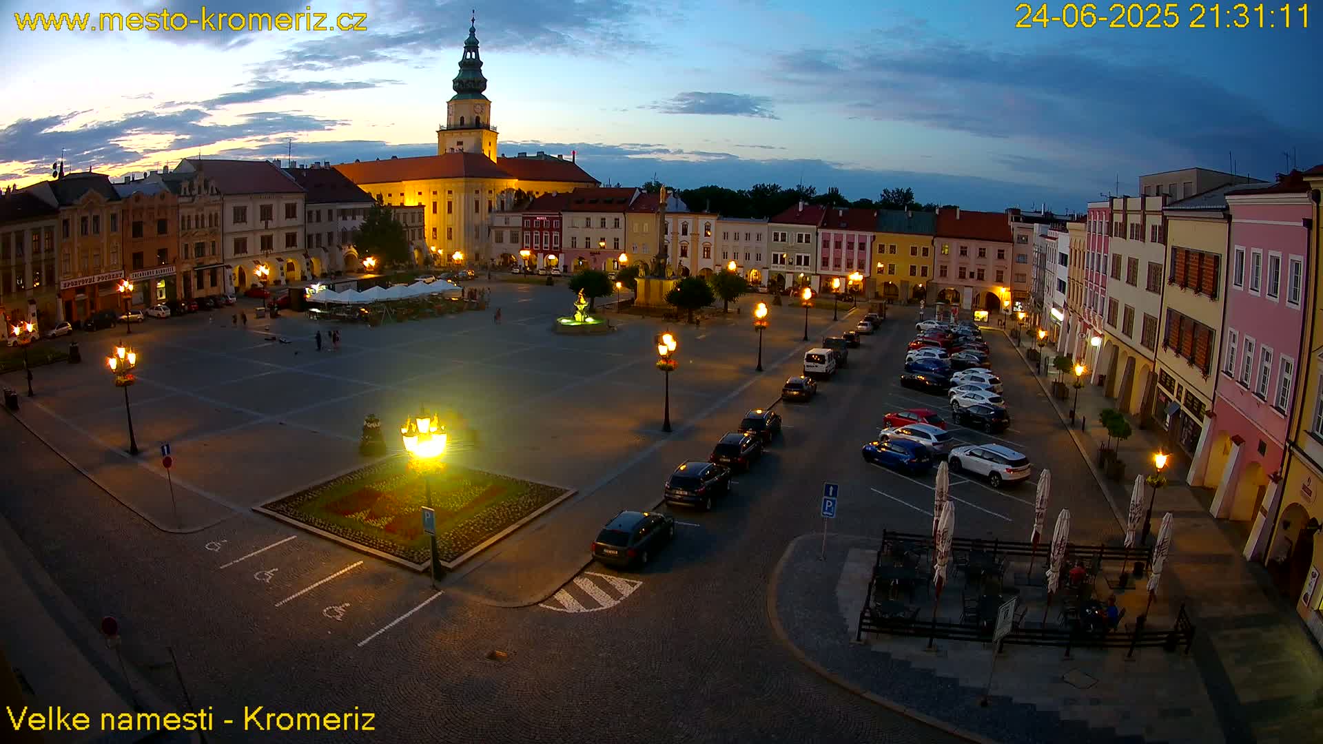A town square at night, illuminated by streetlights, with cars parked along the perimeter and a few people sitting at outdoor tables.