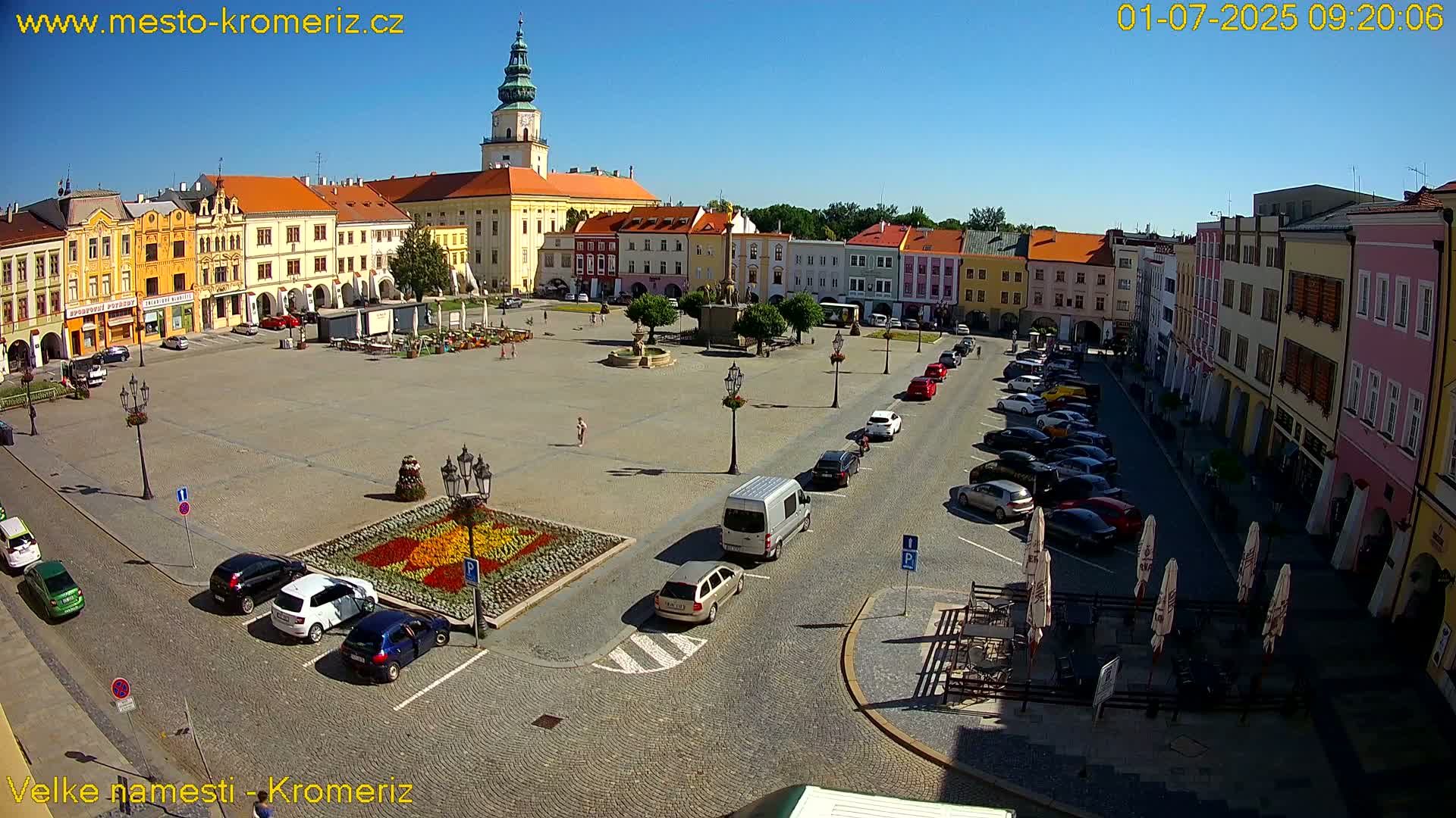 A large town square with colorful buildings surrounding it, cars parked along the sides, and a flower garden in the center, under sunny conditions.